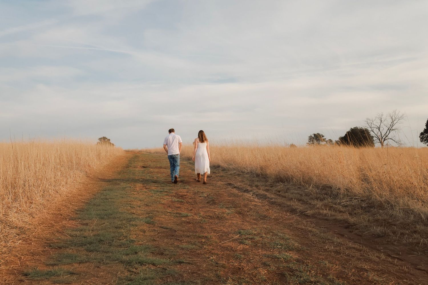 Couple holding hands walking along a dirt path through golden wheat fields under a cloudy sky.