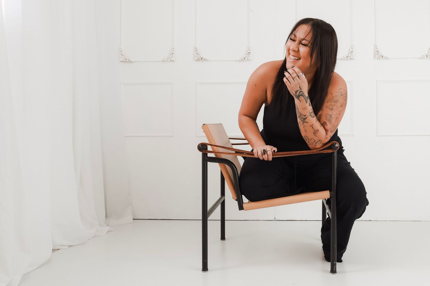 Woman in black outfit sitting on modern chair, smiling with tattooed arms, in bright white studio setting.