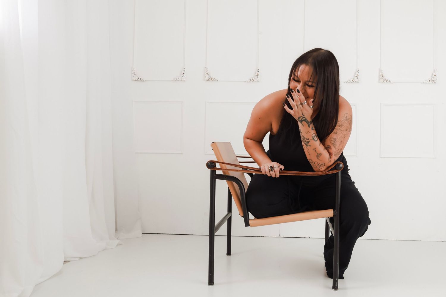 Woman in black outfit sitting on modern chair, hand over mouth, against white wall background in a studio