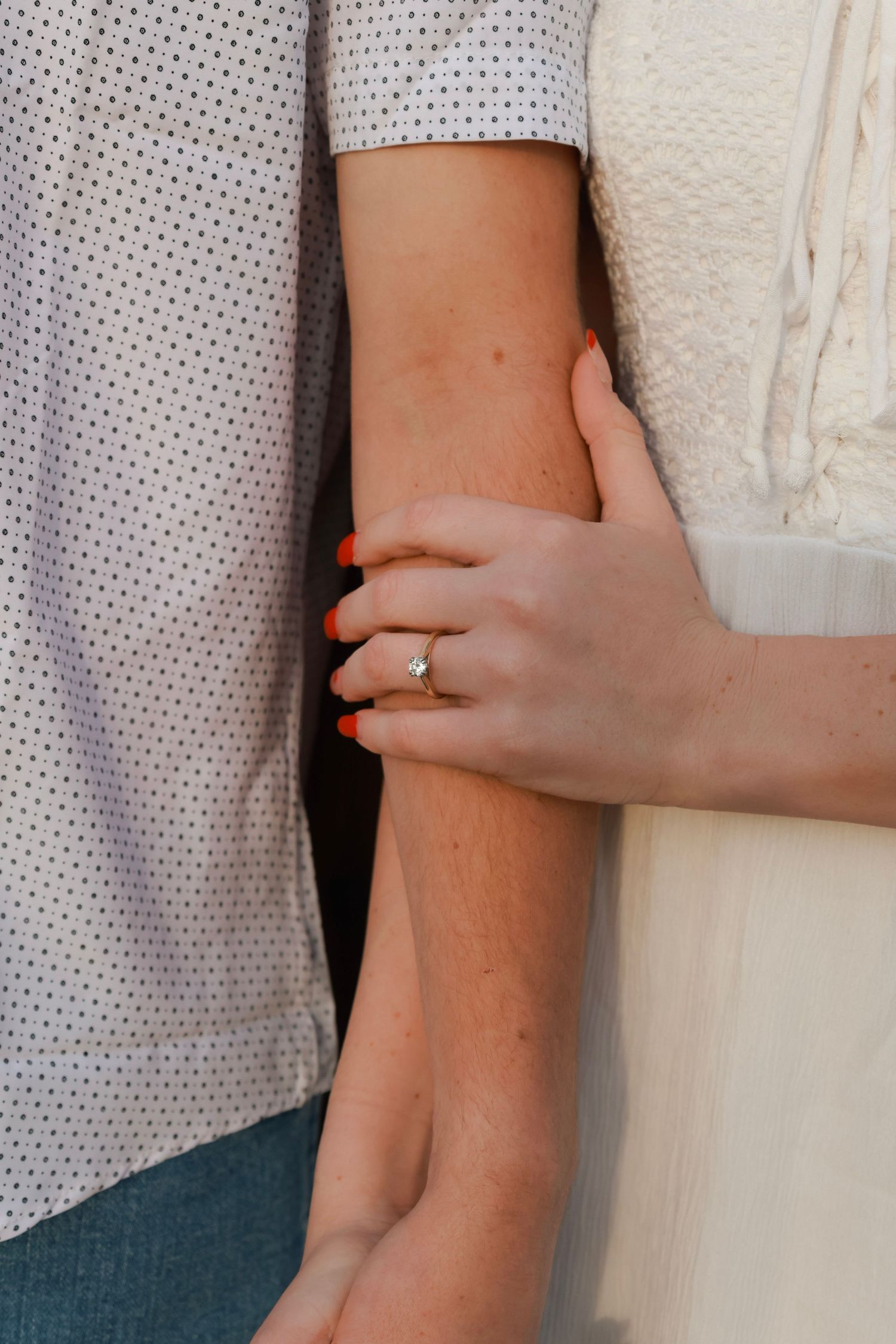 Close-up of a hand with red nails and a gold engagement ring gently holding another person's arm.