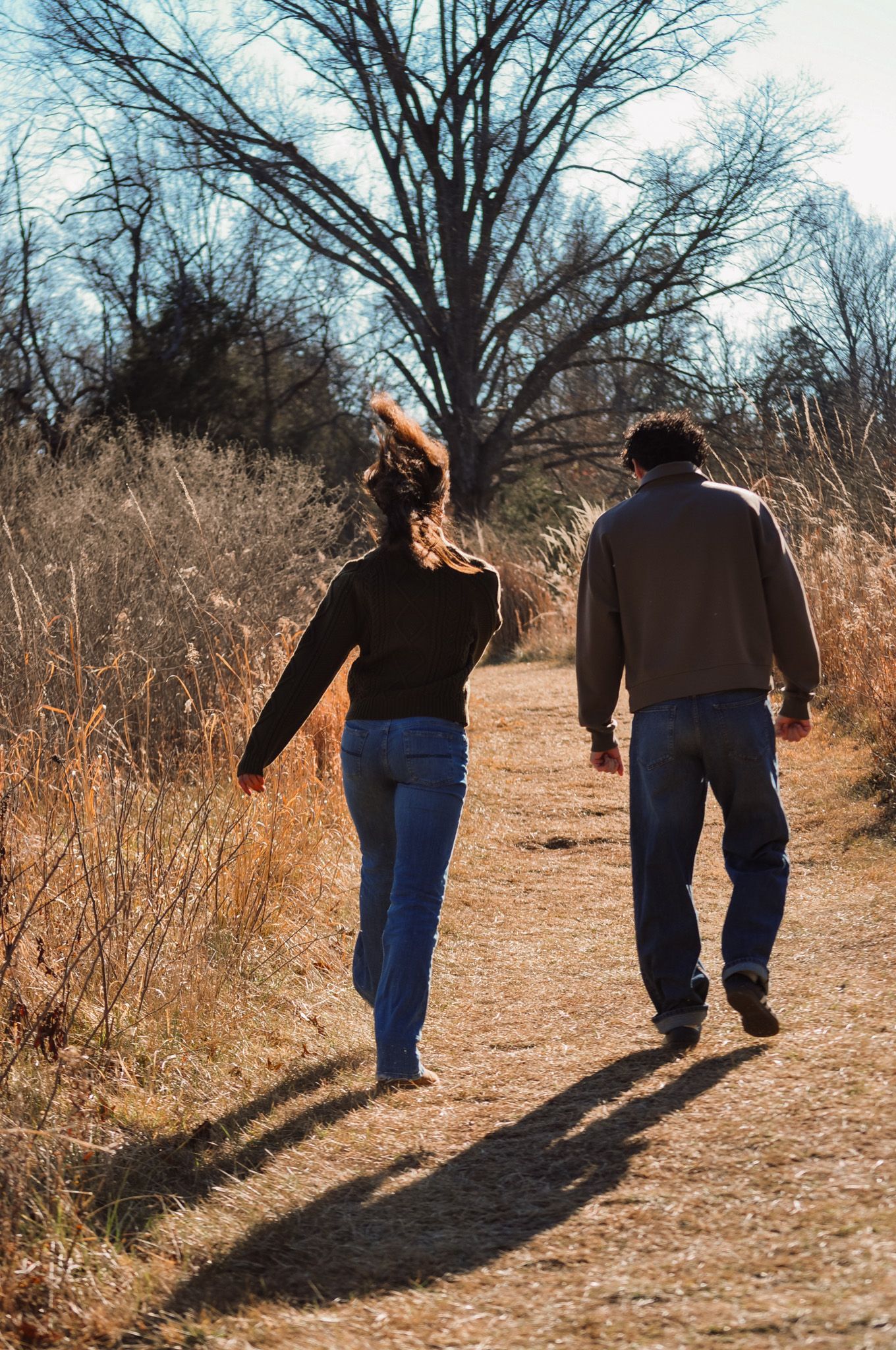 Couple walking hand in hand on a dirt path through dry autumn fields with bare trees in the background.