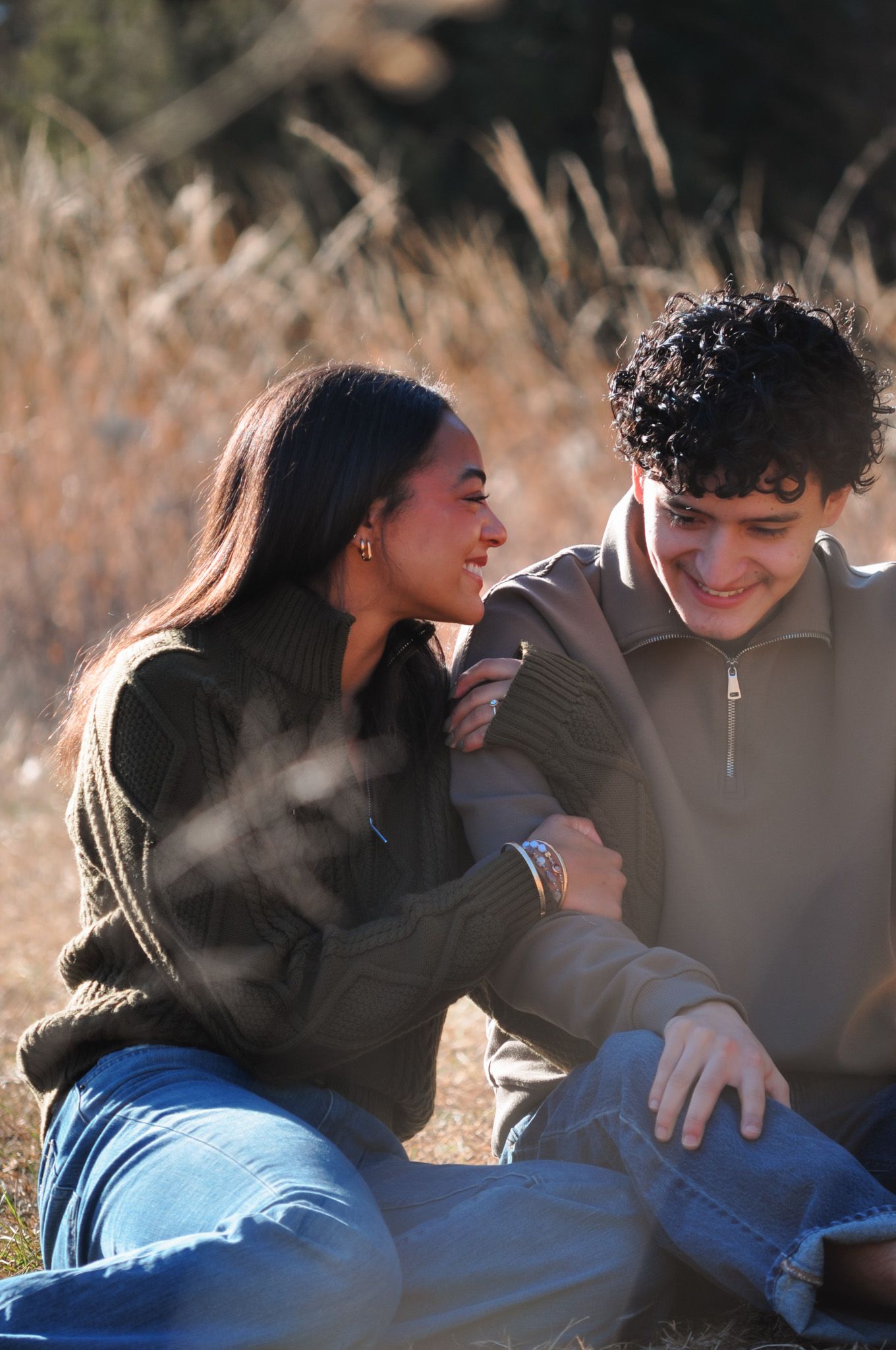 A smiling couple sits together outdoors in a sunny field, sharing a warm and affectionate moment.