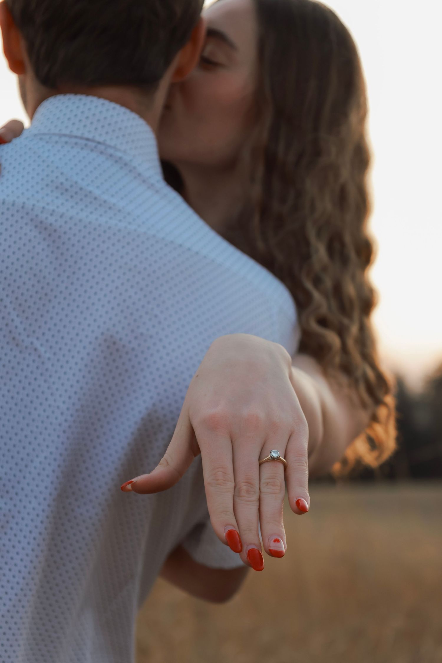 Couple sharing a kiss at sunset while woman displays her engagement ring with red nails toward the camera.