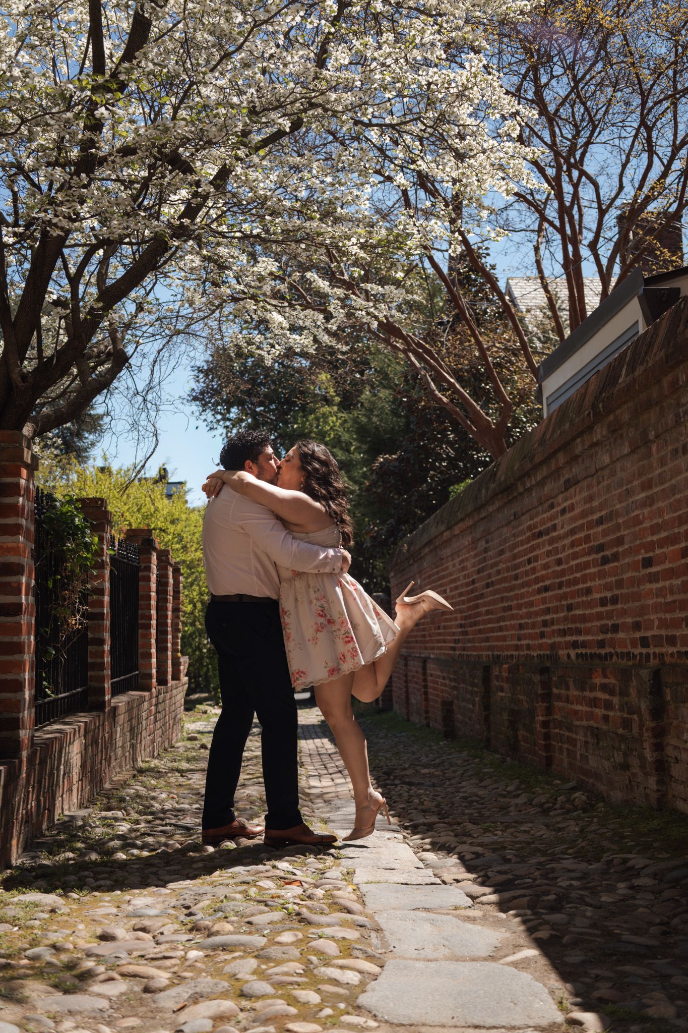 Couple sharing a romantic kiss on a cobblestone path under blooming white cherry trees beside a brick wall.