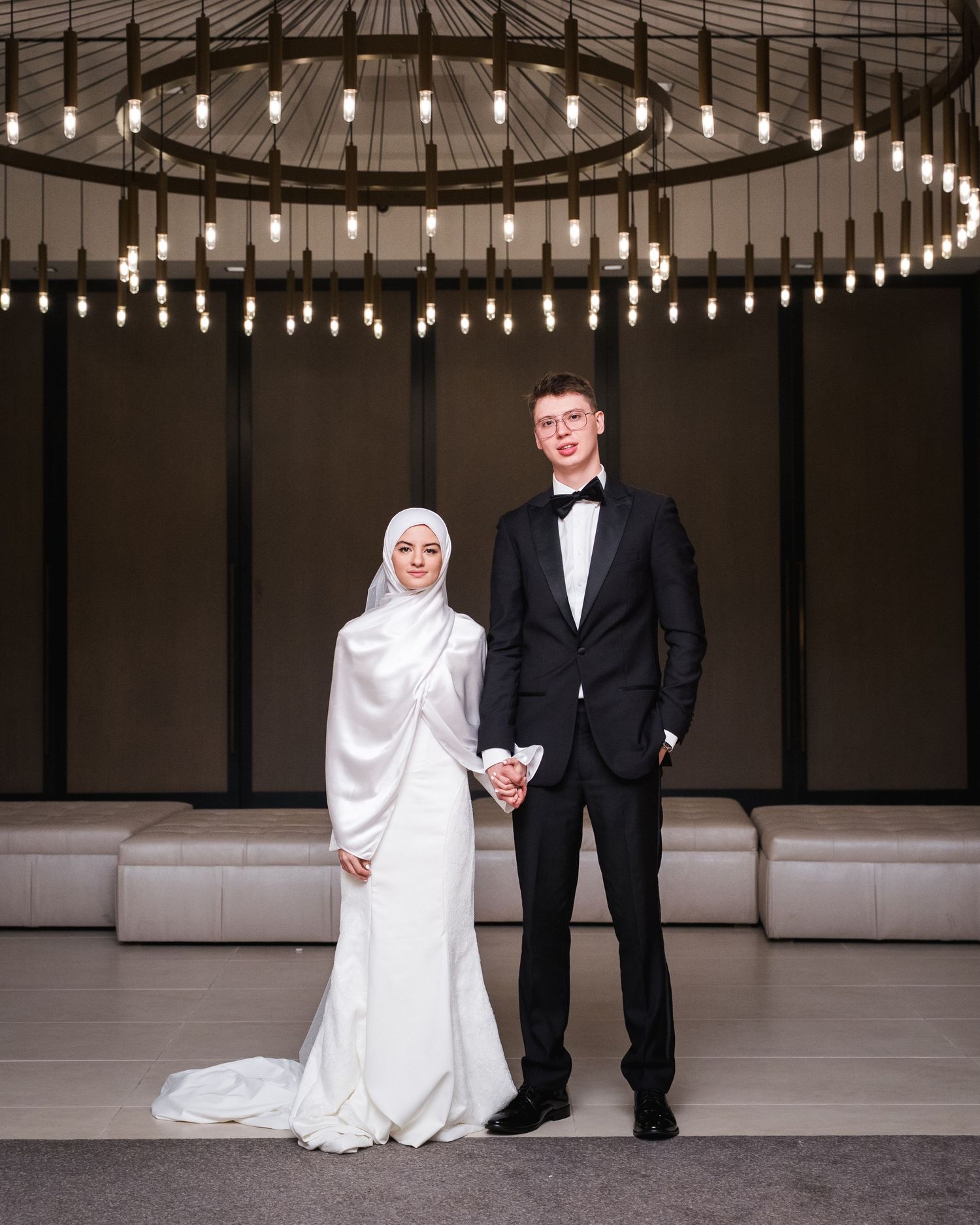 Elegant wedding couple posing under circular chandelier, bride in white hijab gown, groom in black tuxedo with bow tie.