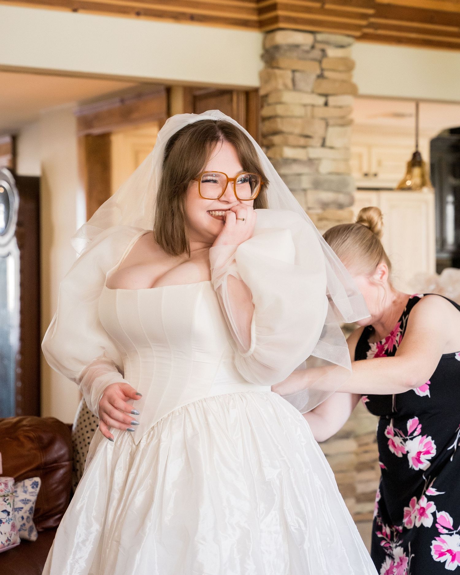 Bride in white ball gown and veil with glasses smiles while bridesmaid adjusts her dress in rustic venue.