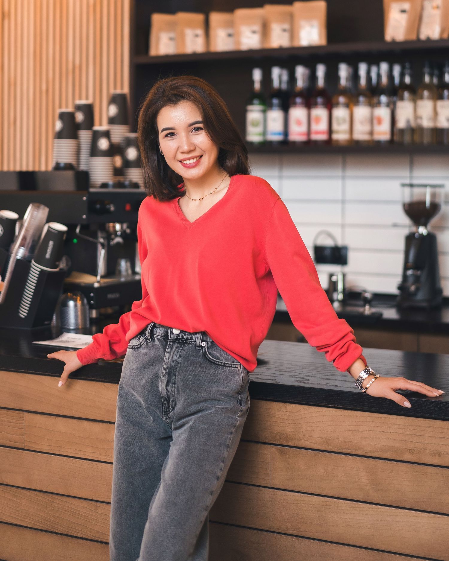 Person in red sweater and jeans posing against wooden coffee bar counter.