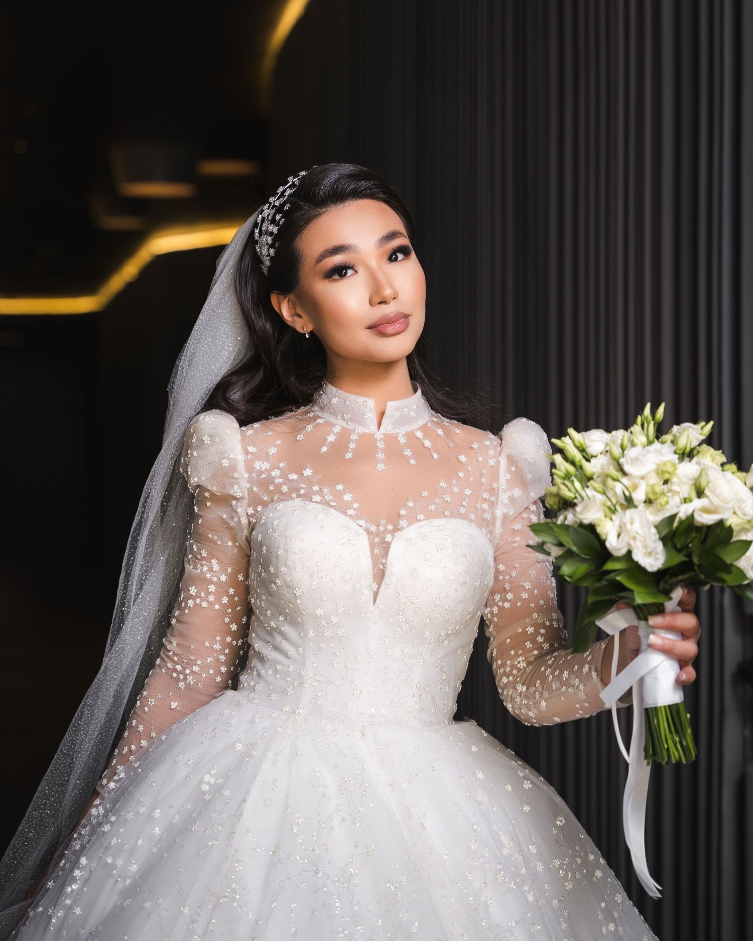 Elegant bride in a pearl-embellished high-neck ballgown with puff sleeves, veil, and white rose bouquet.
