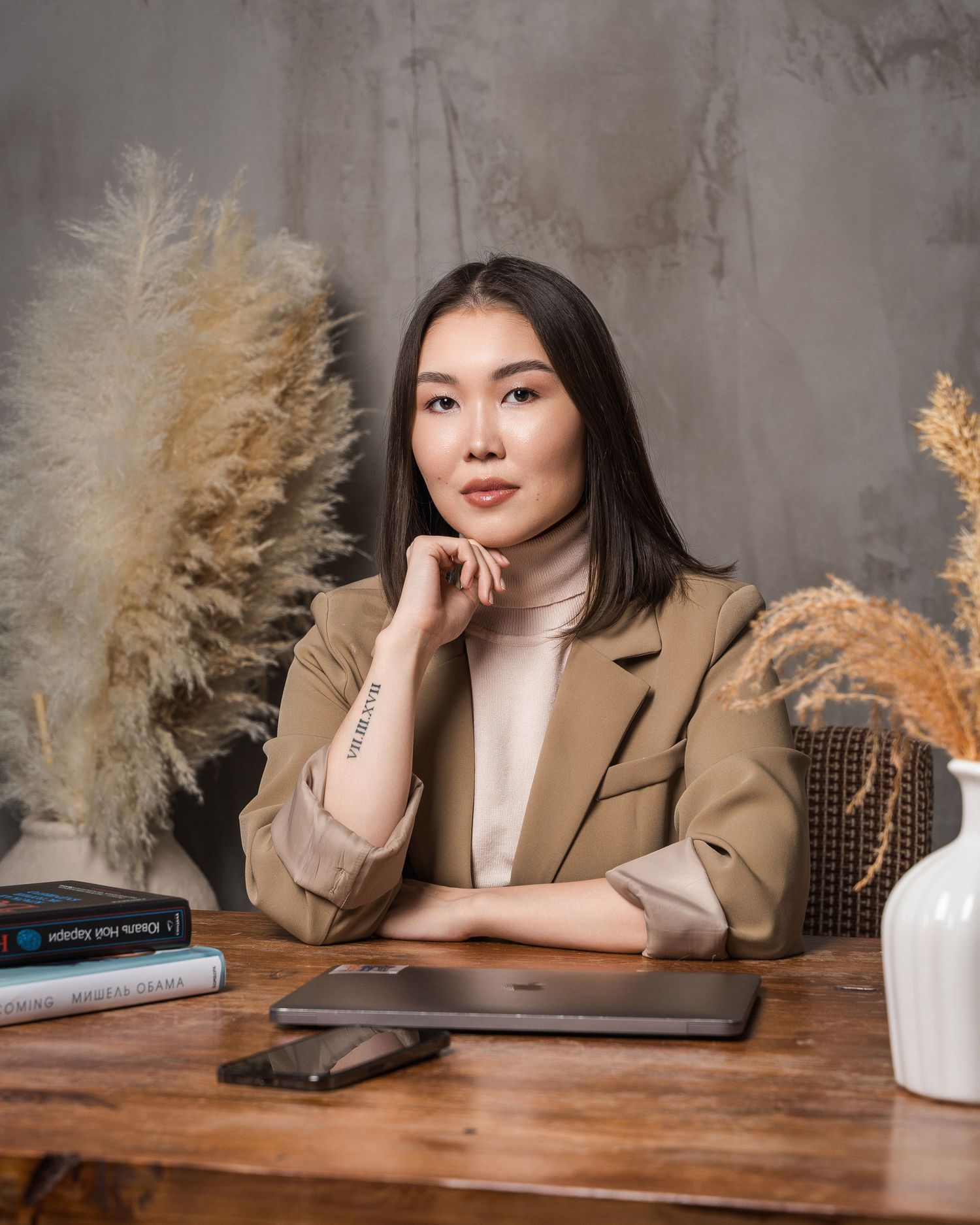 Professional portrait in tan blazer sitting at wooden desk with pampas grass decor.