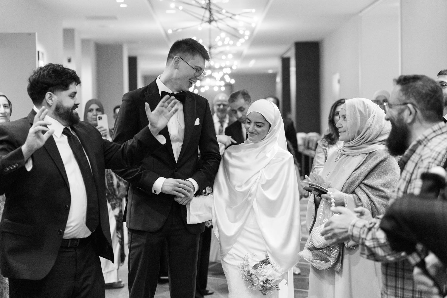 Black and white photo of a Muslim bride in hijab walking with groom in tuxedo at elegant wedding reception.
