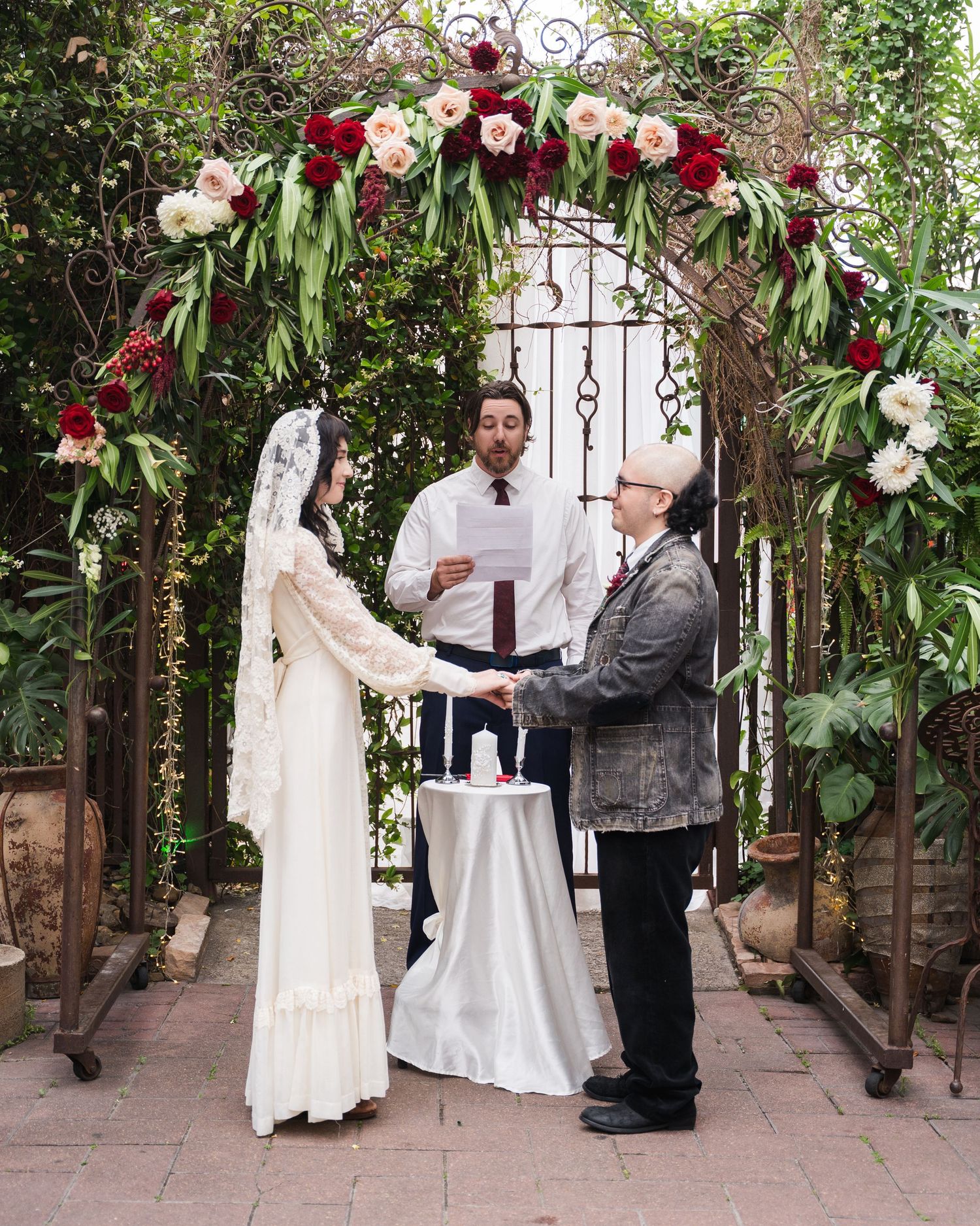 Couple exchanges vows under floral arch with red and white roses at outdoor garden wedding ceremony.