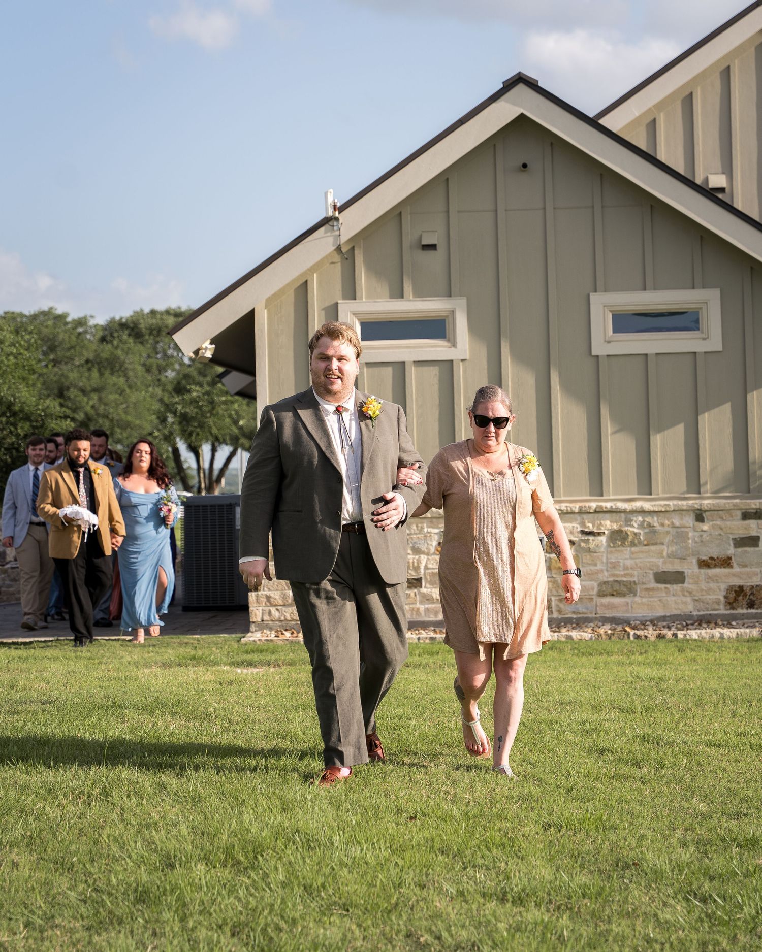 A man in a gray suit escorts a woman in a peach dress across a green lawn at an outdoor wedding venue.