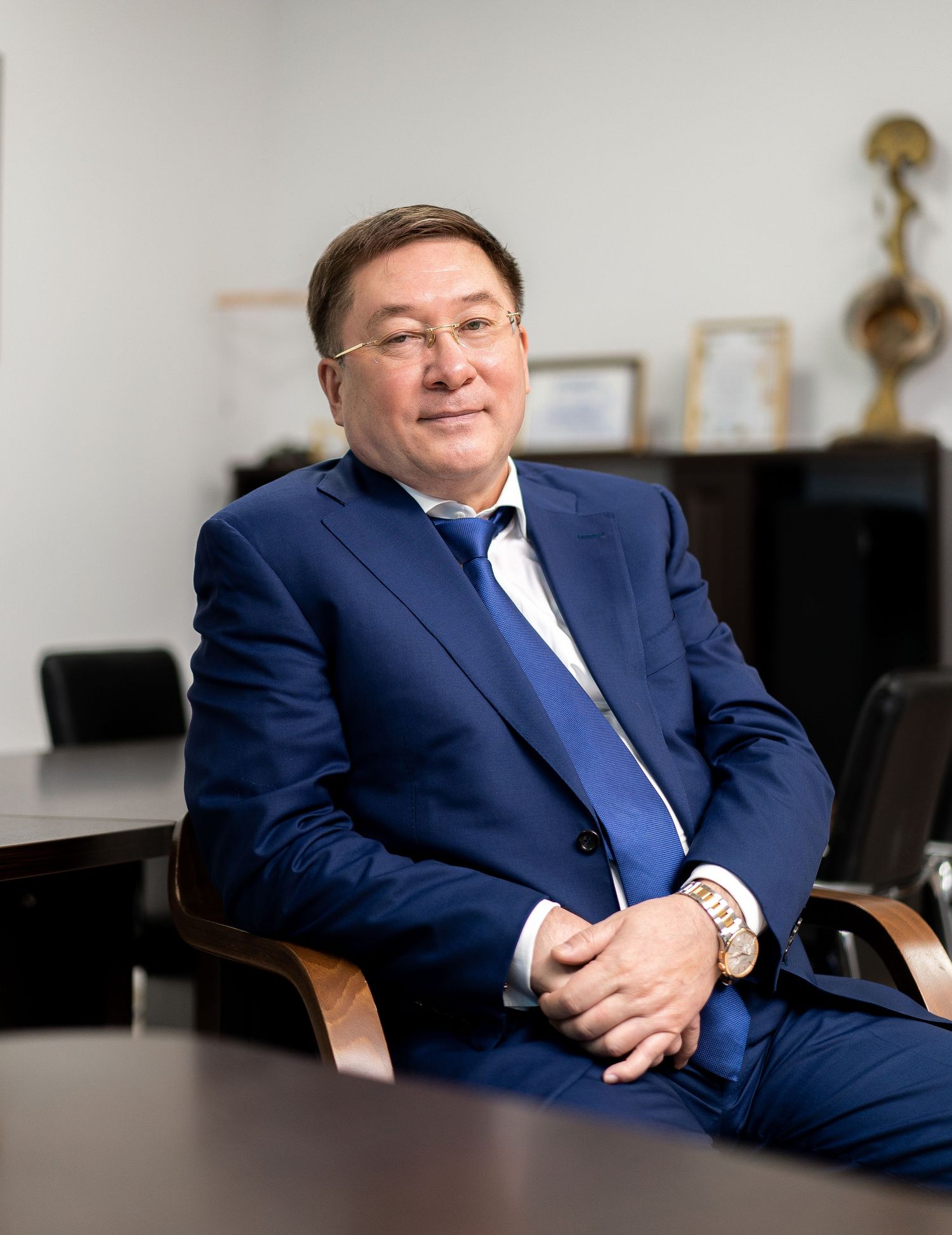 Business executive in navy suit seated in office with awards visible in background.