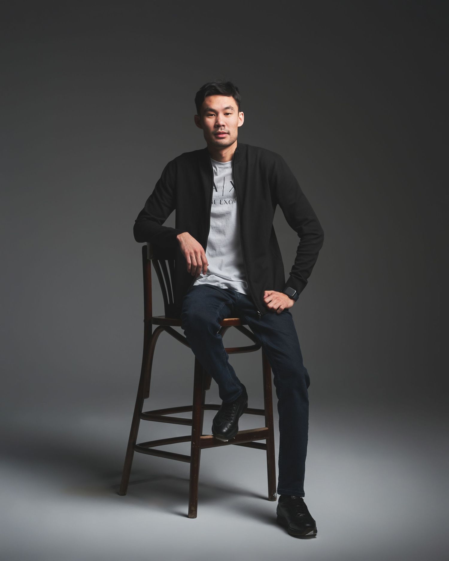 Person in casual attire posing with wooden chair in studio setting with dramatic lighting.