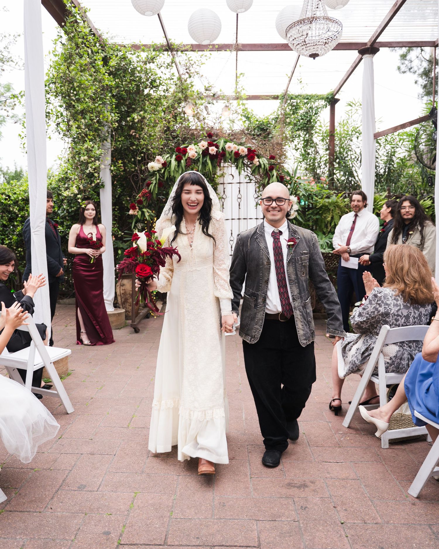 Newlyweds walk down outdoor aisle holding hands after wedding ceremony under floral arch with guests.