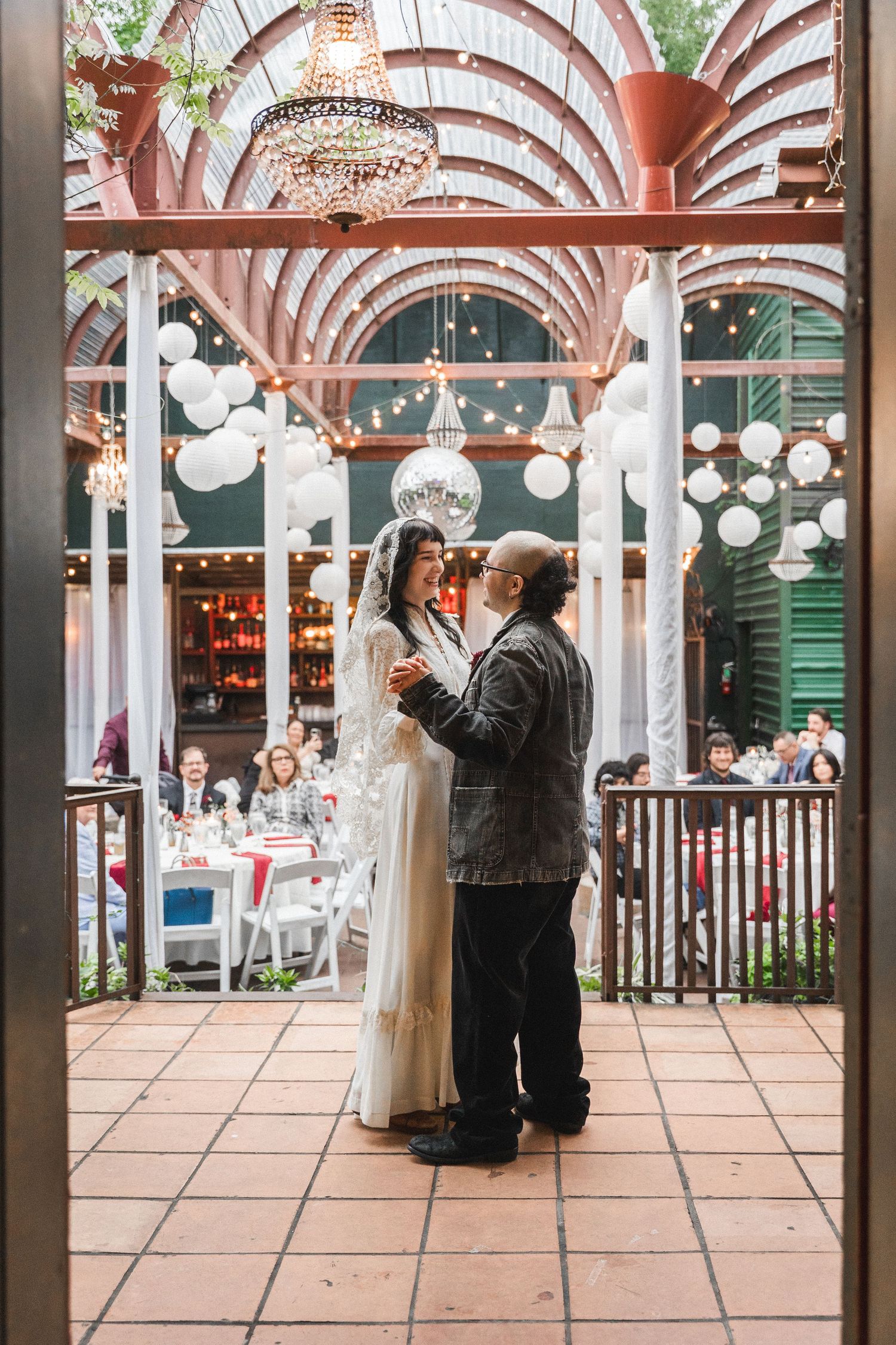 Bride and groom share first dance in elegant venue with chandeliers, white lanterns, and guests seated at decorated tables.