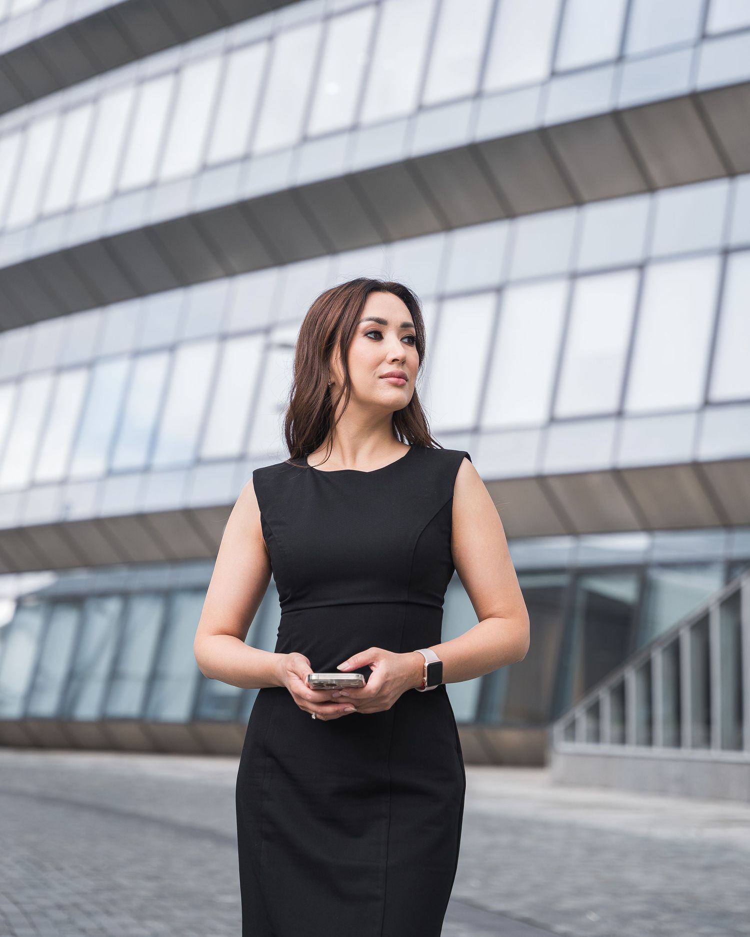 Professional in black sleeveless dress poses near modern glass building exterior.