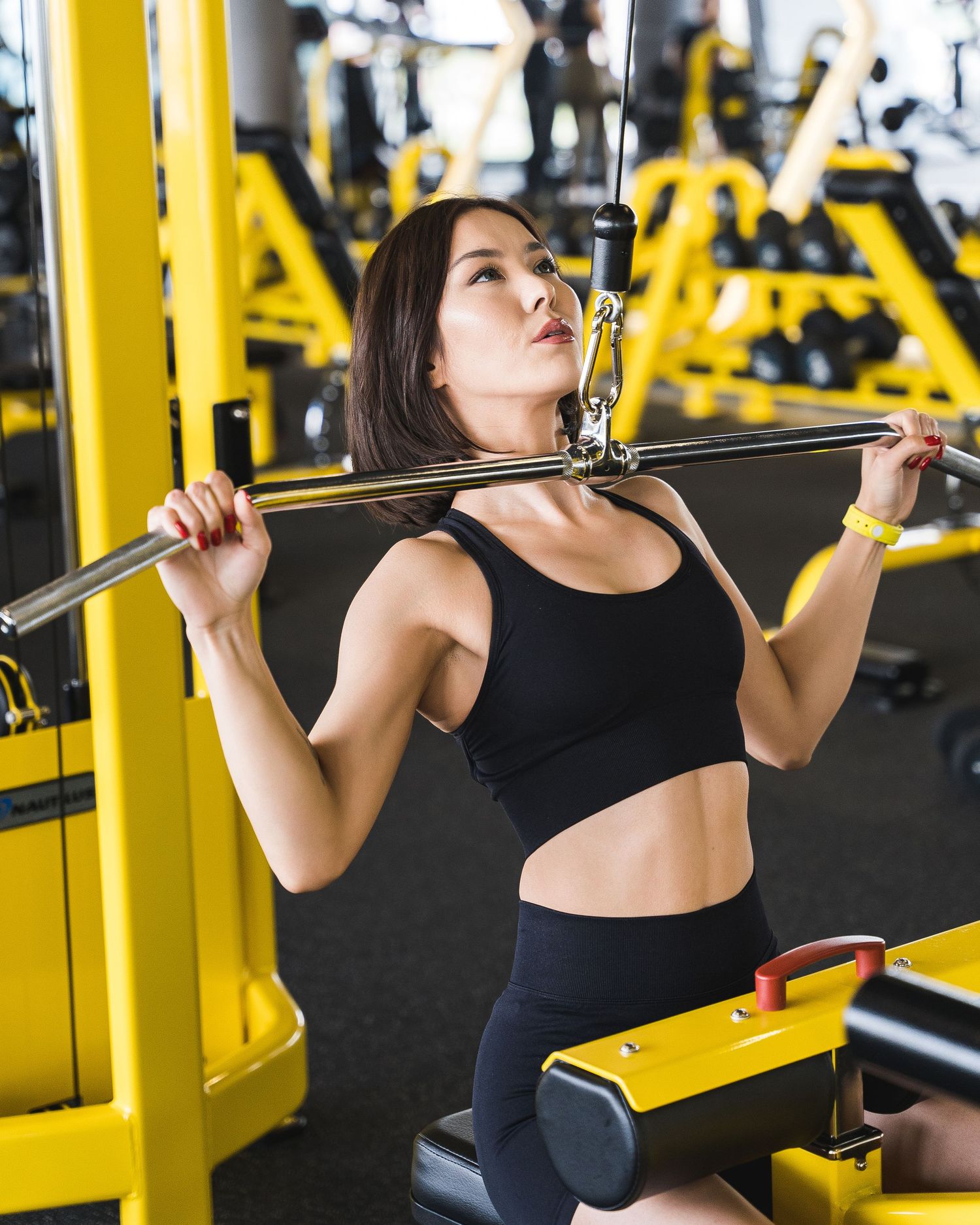 Woman in black athletic wear performing lat pulldown exercise at a gym with yellow equipment.