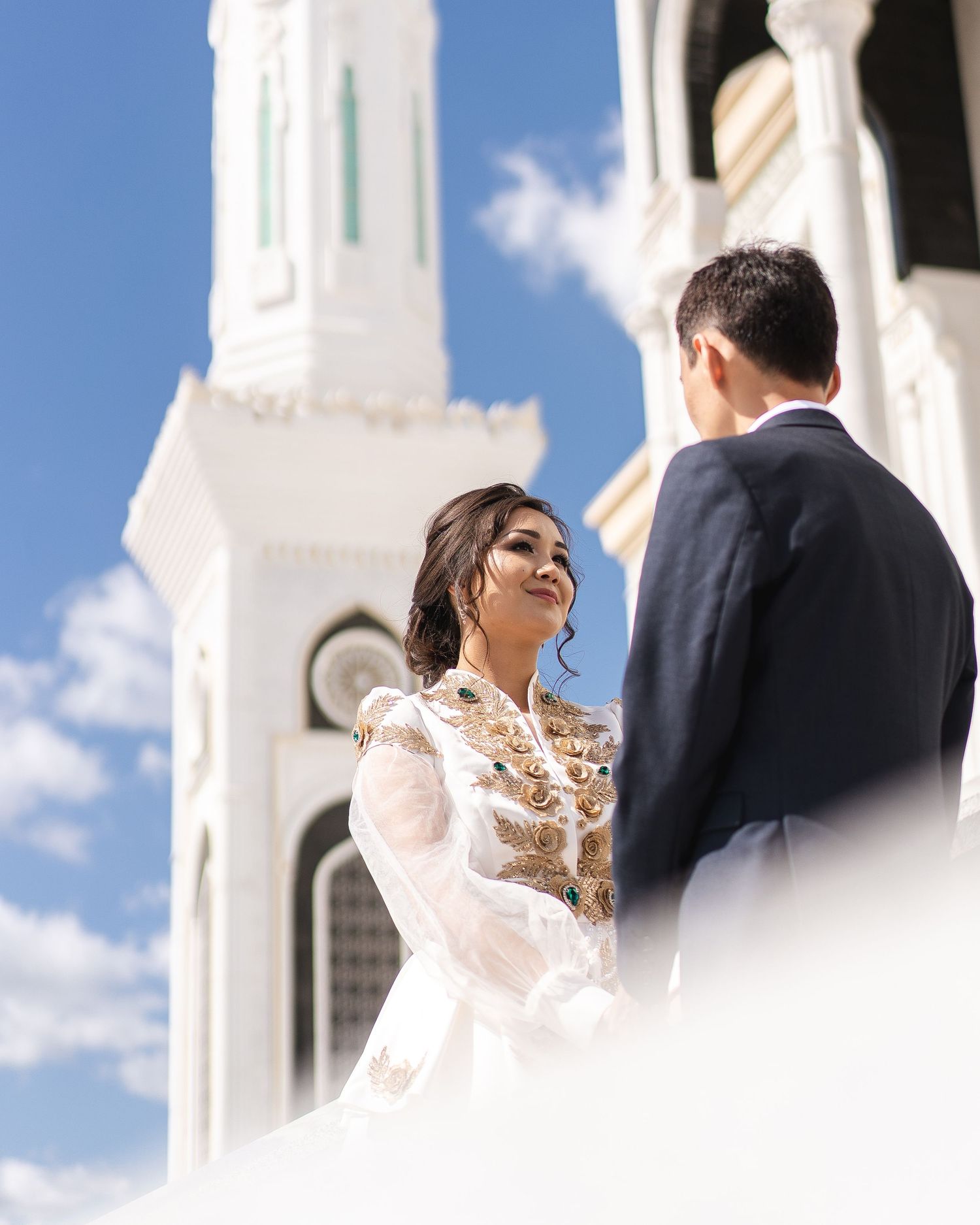 Bride in ornate white lace gown with gold embroidery stands before a white mosque minaret under blue sky.