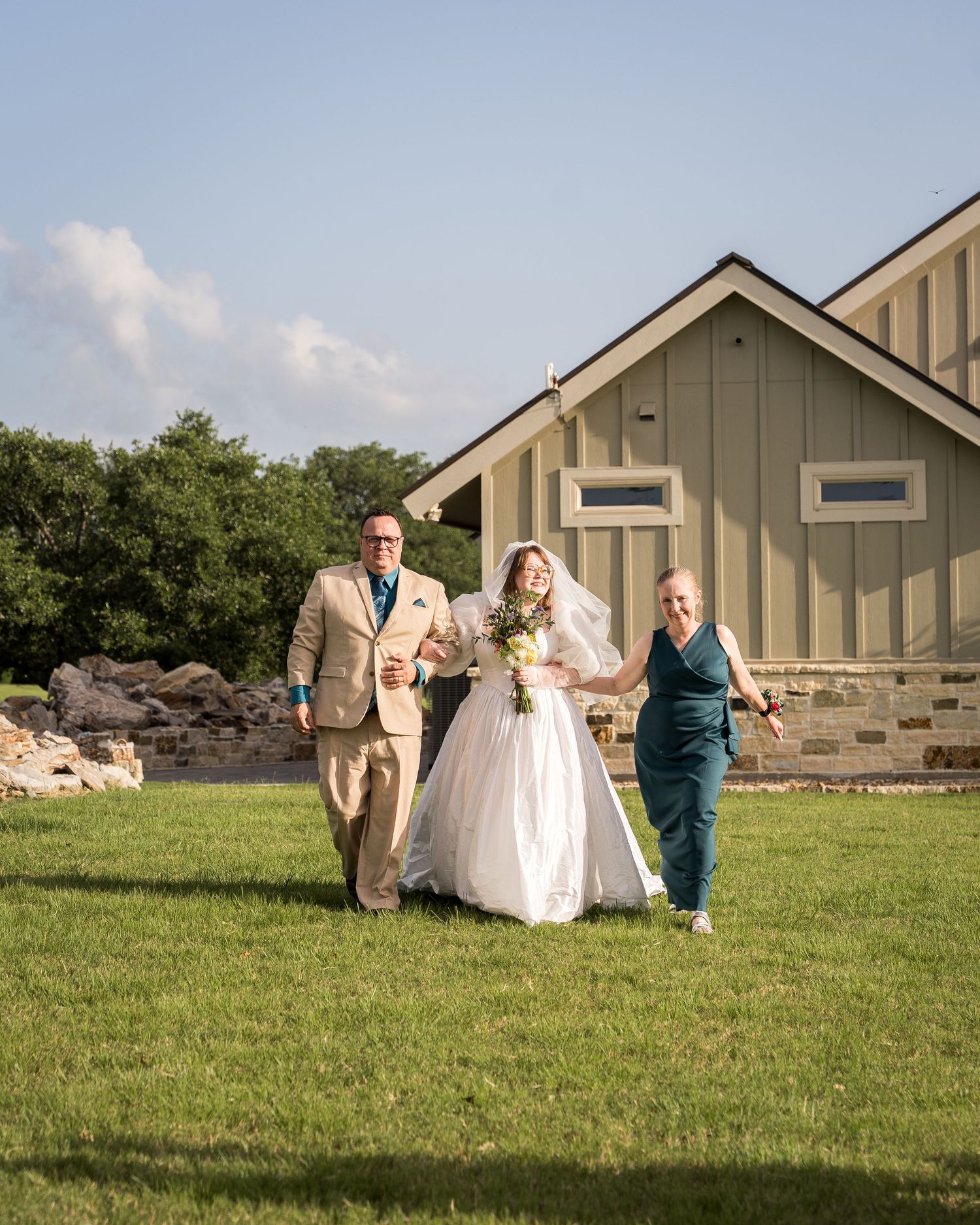 A bride in a white gown and veil walks with two escorts across a sunny lawn toward an outdoor wedding ceremony.