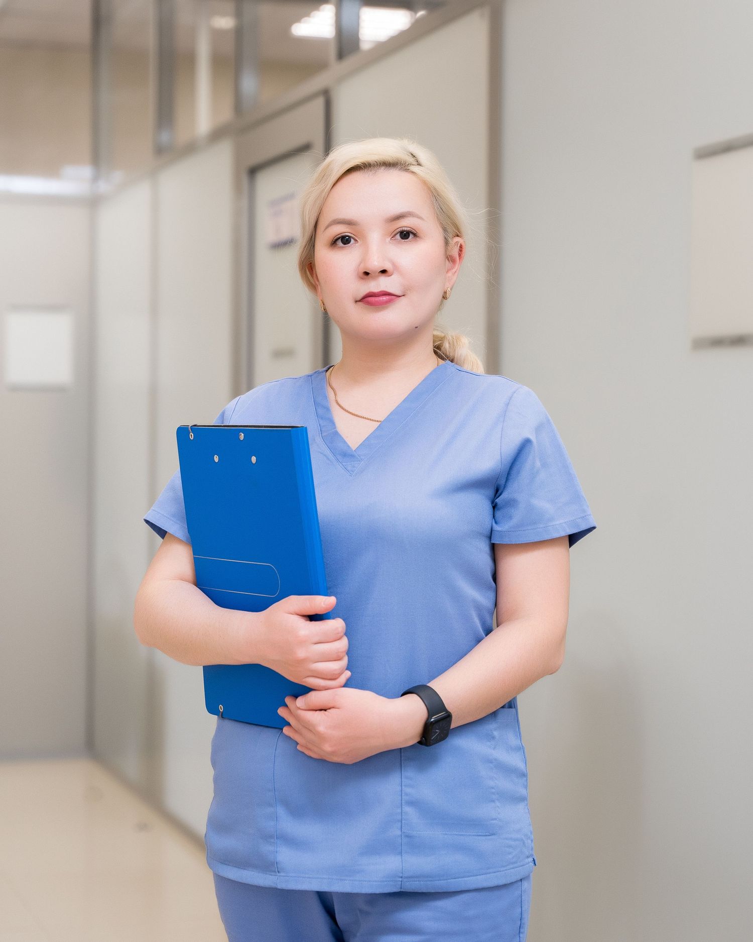 Healthcare professional in light blue scrubs holding a blue clipboard in a medical hallway.