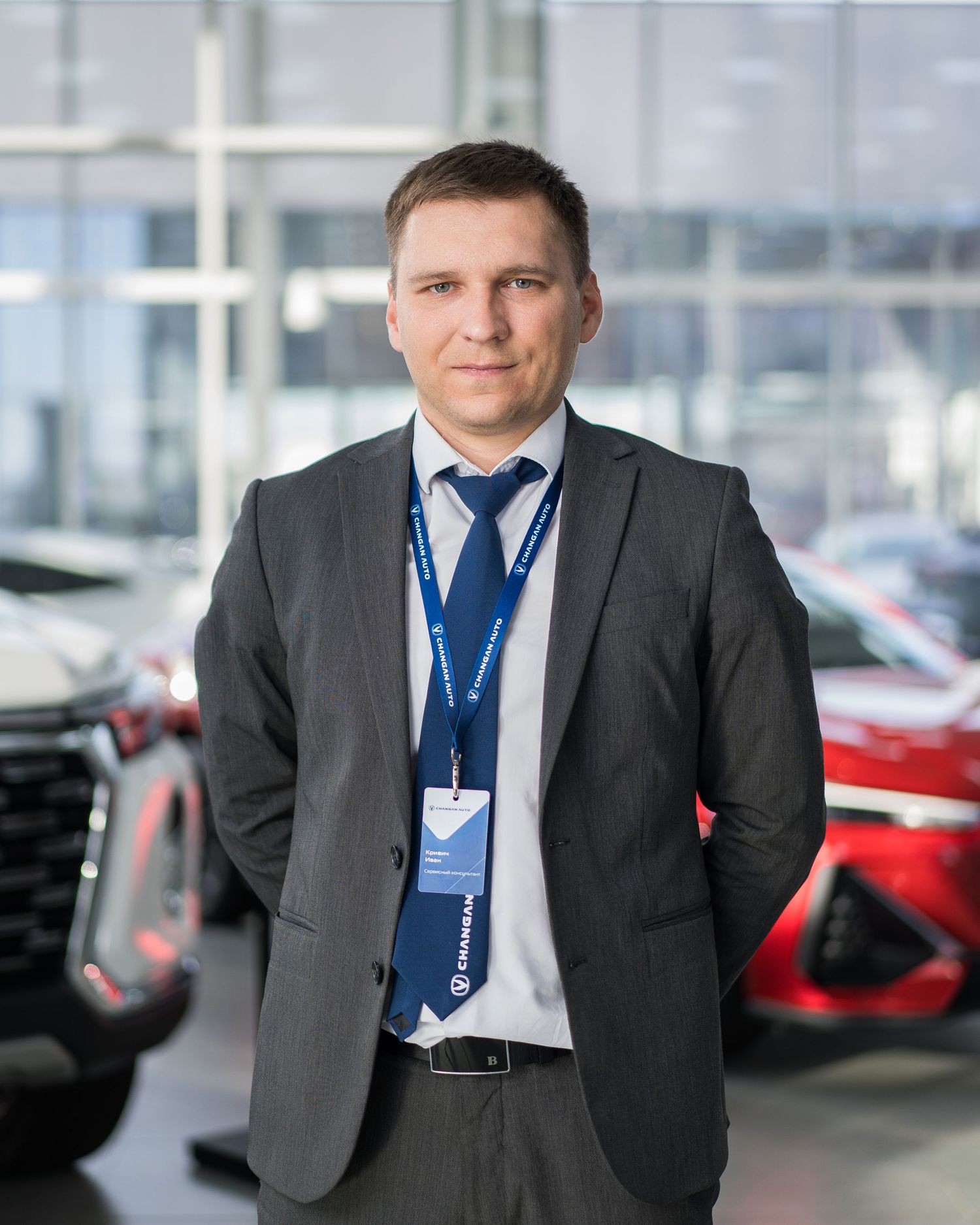 Professional in grey suit and blue tie standing in car dealership showroom.