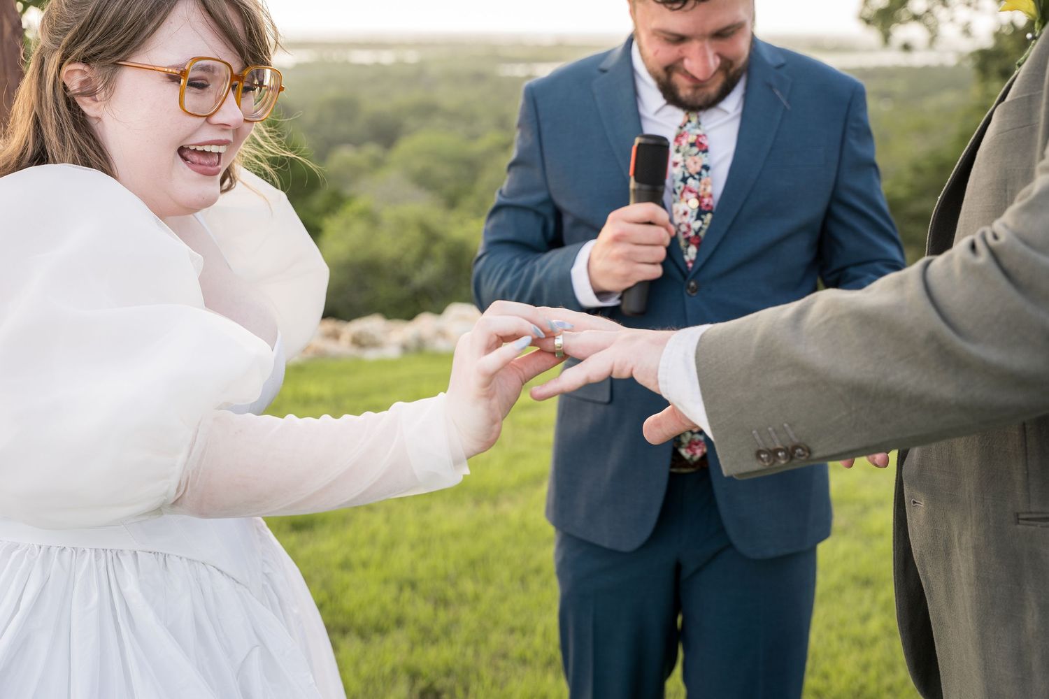 Bride laughing joyfully as groom places wedding ring on her finger during outdoor ceremony.