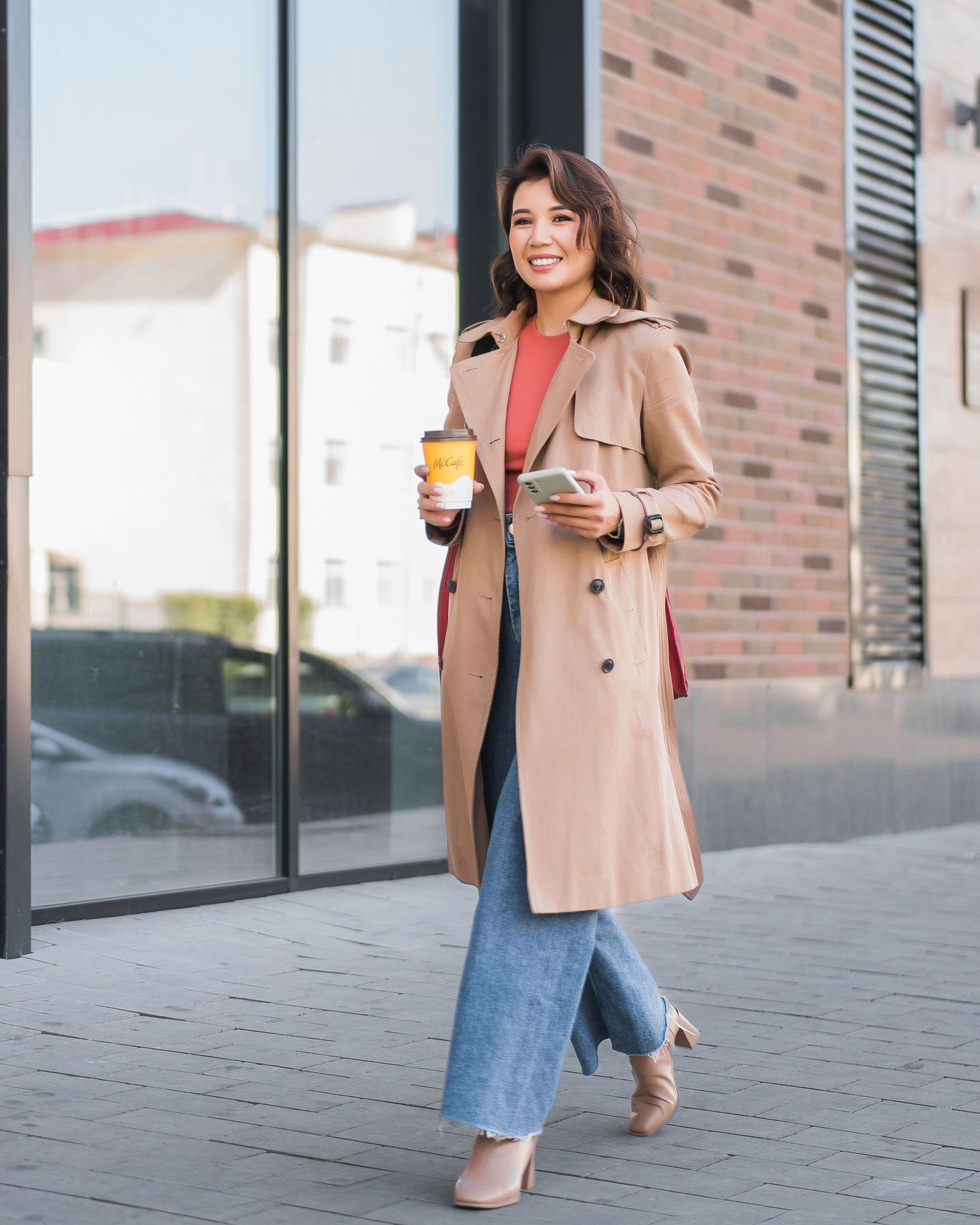 Stylish person in beige blazer and blue dress walking outdoors near modern building with coffee cup.