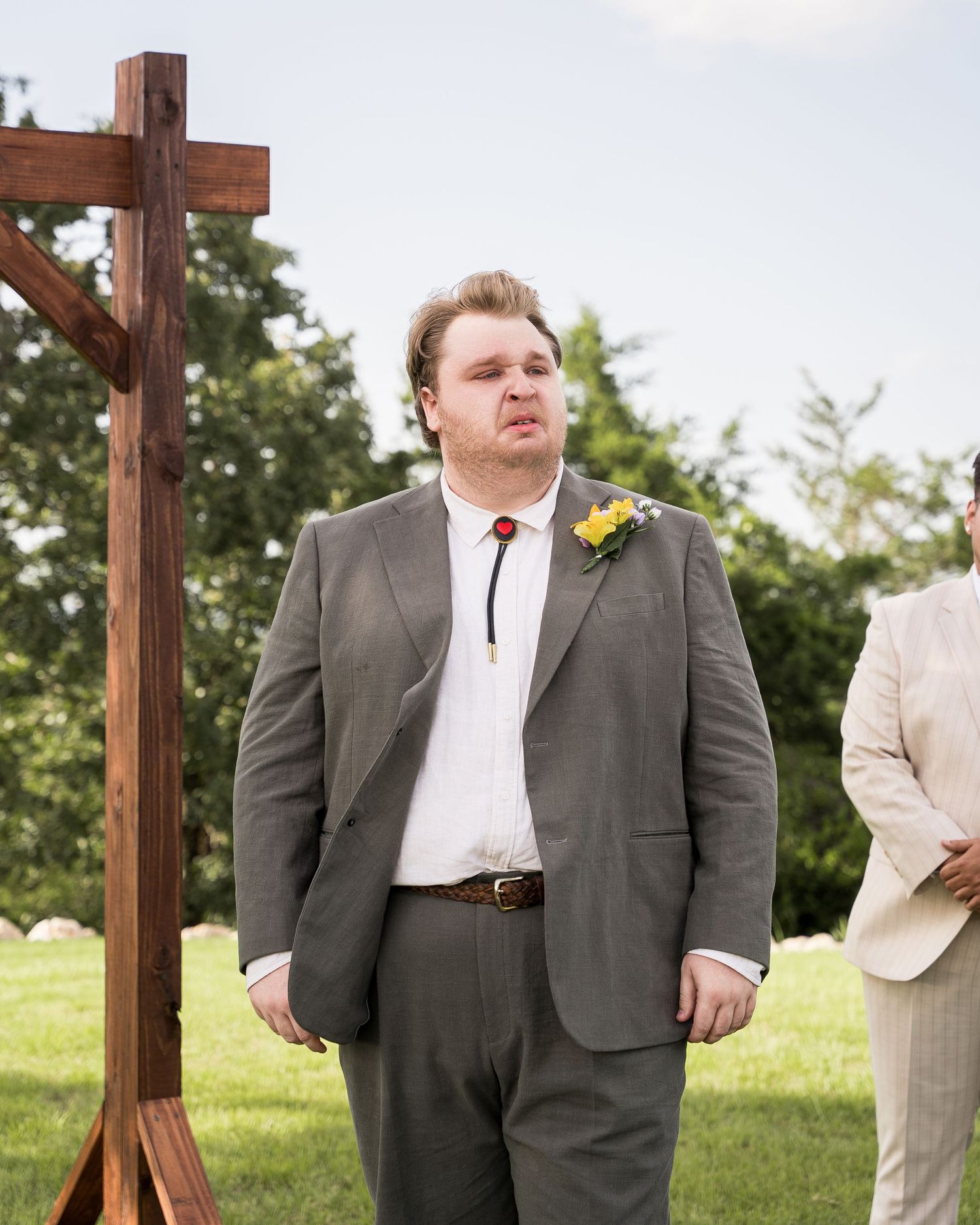 Groom in gray suit with yellow boutonniere stands at outdoor wedding ceremony beside wooden cross.