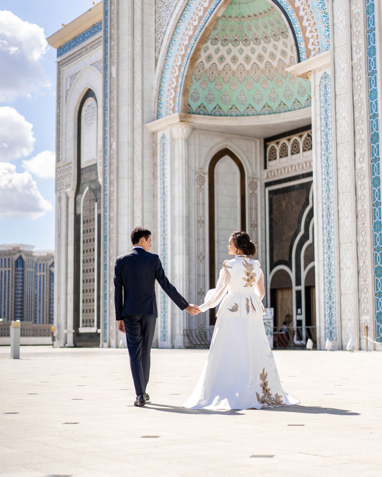 Bride and groom holding hands walking toward ornate mosque with turquoise dome on sunny wedding day.
