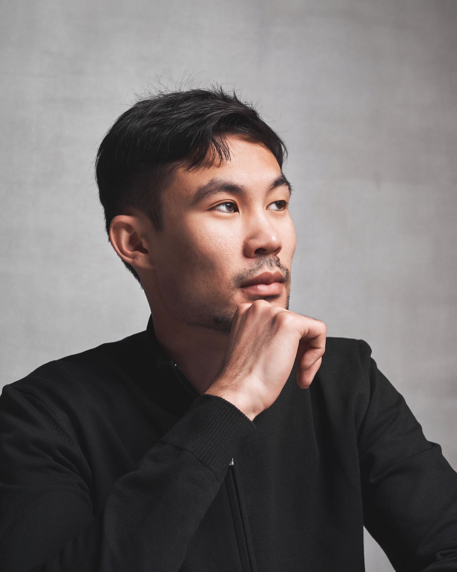 Person in black jacket photographed in dramatic profile against grey background.