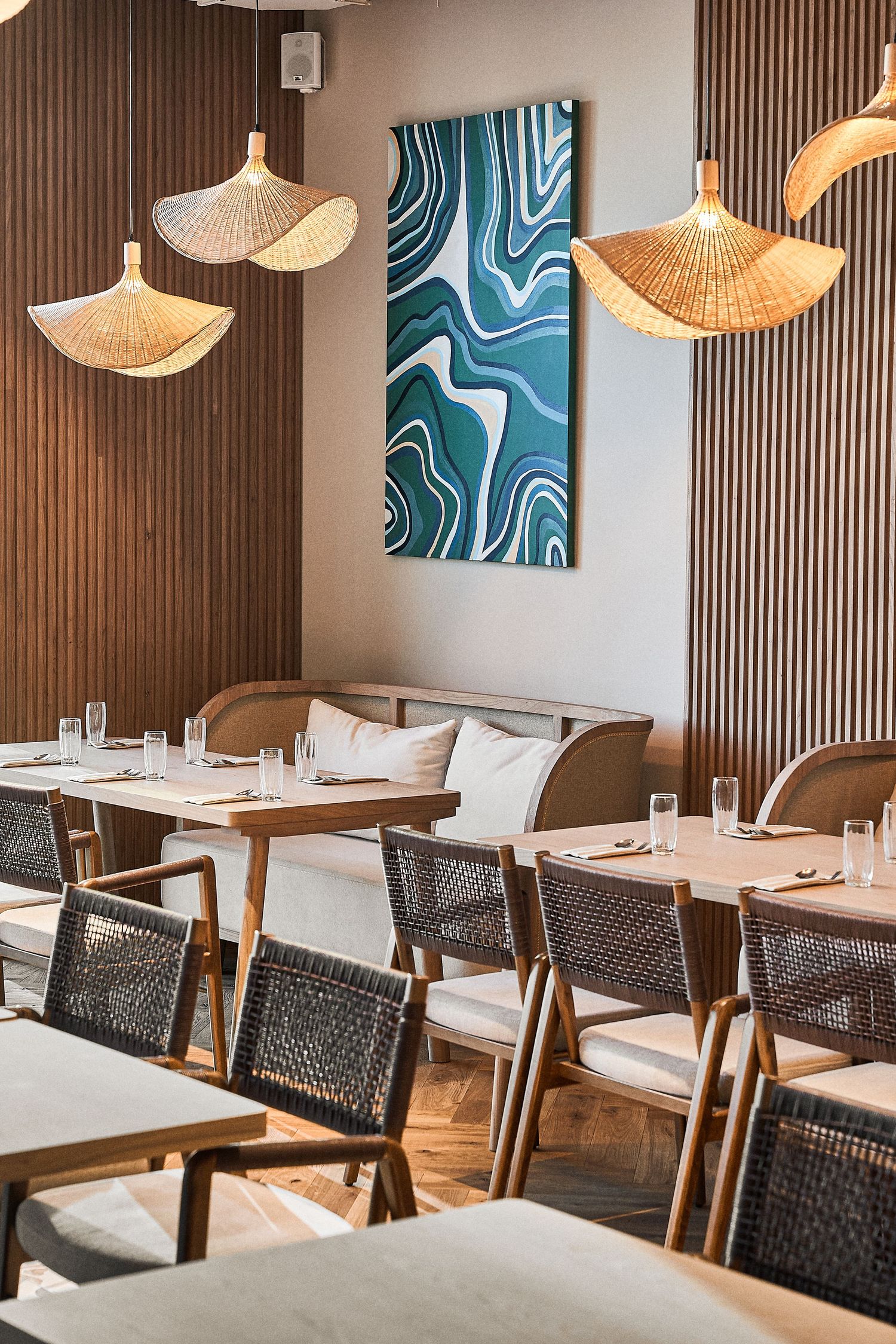 Restaurant dining room with blue art panels, woven pendant lights, and modern wooden furniture.
