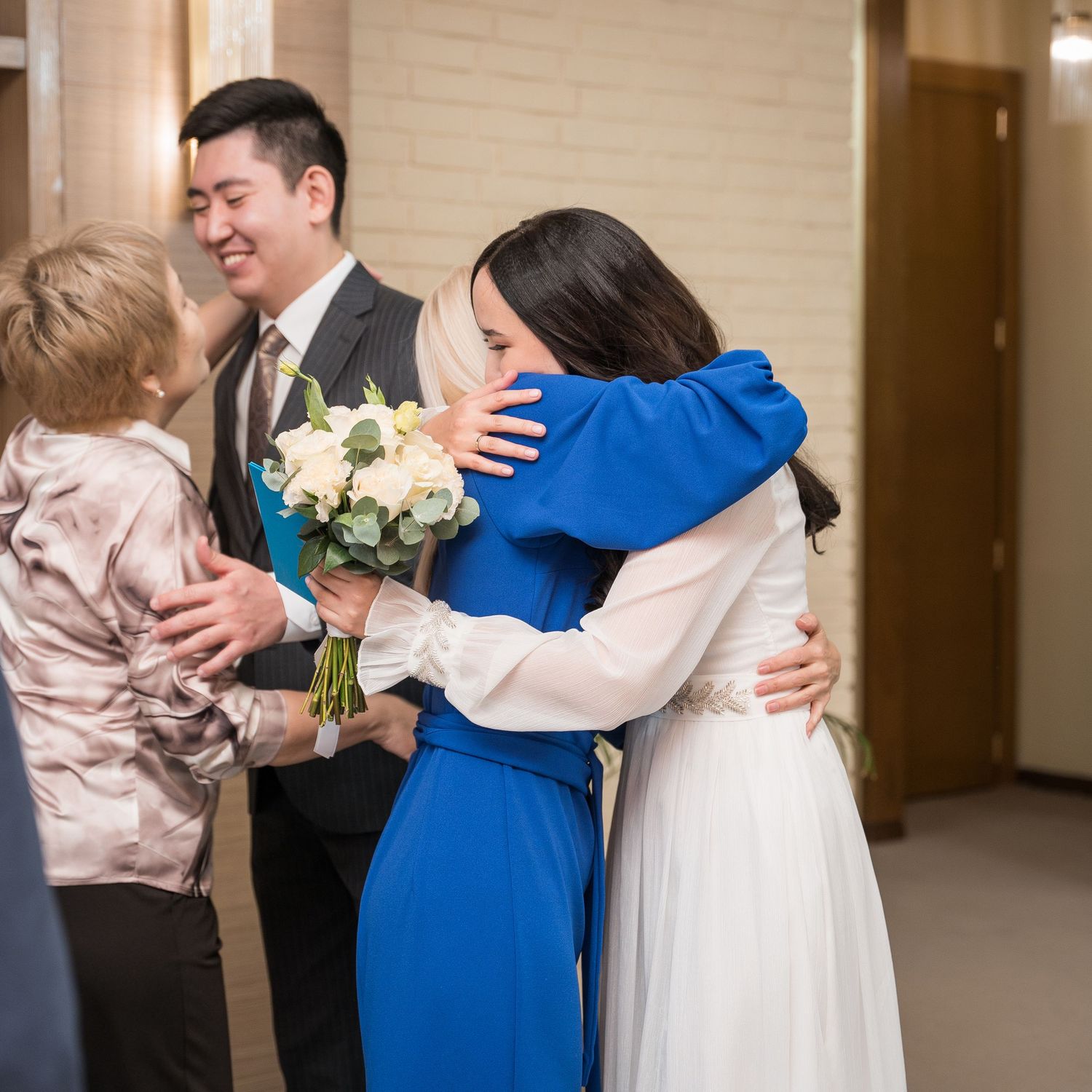 Wedding guests embrace bride in white dress while groom in suit holds bouquet in decorated hallway.