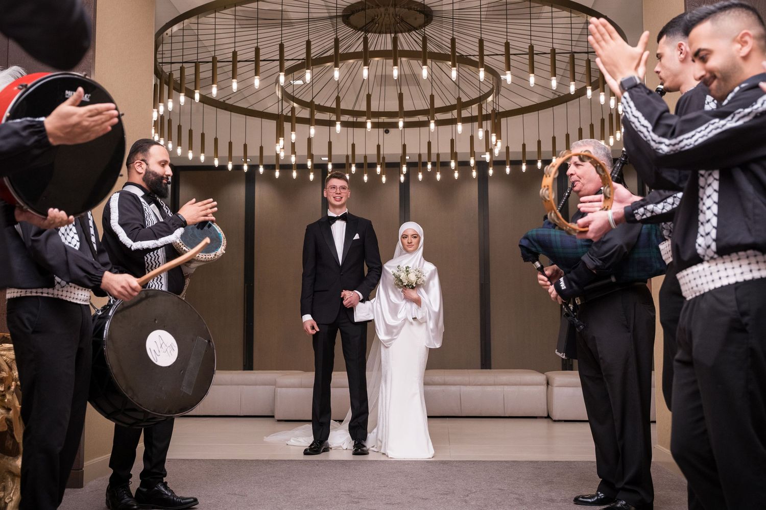 Muslim bride and groom stand together at wedding ceremony with traditional drummers and guests celebrating around them.