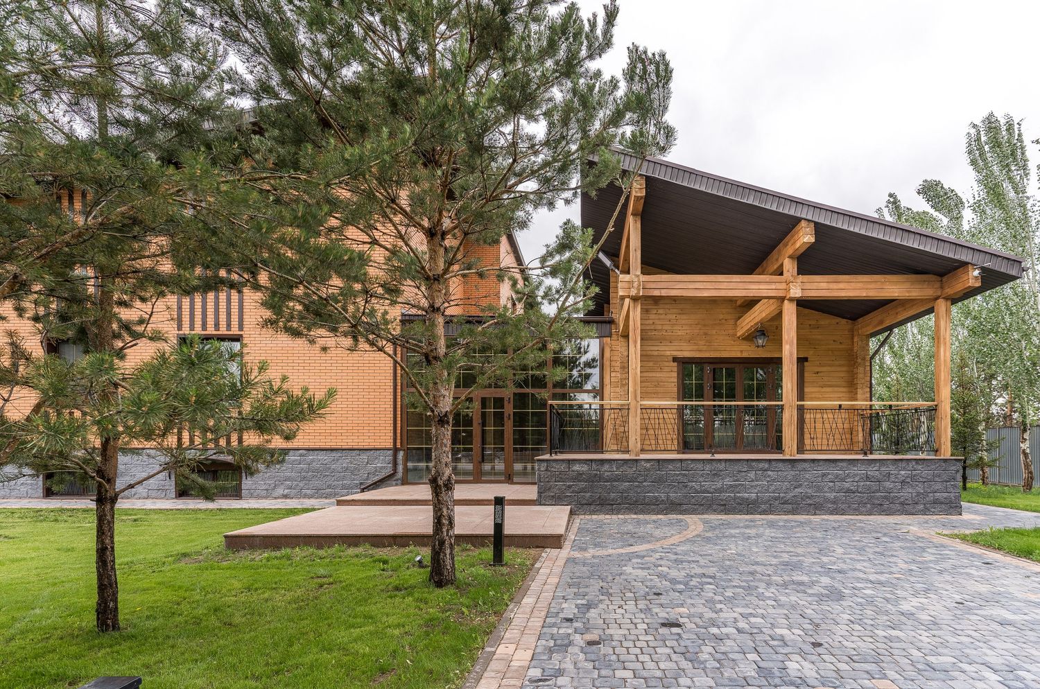 Modern wooden pavilion with stone patio surrounded by pine trees and landscaped grounds.