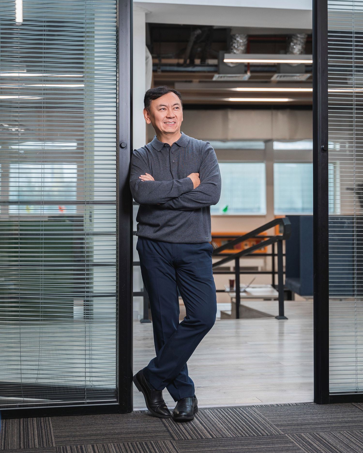 Professional in dark suit leaning against office wall with glass panels.