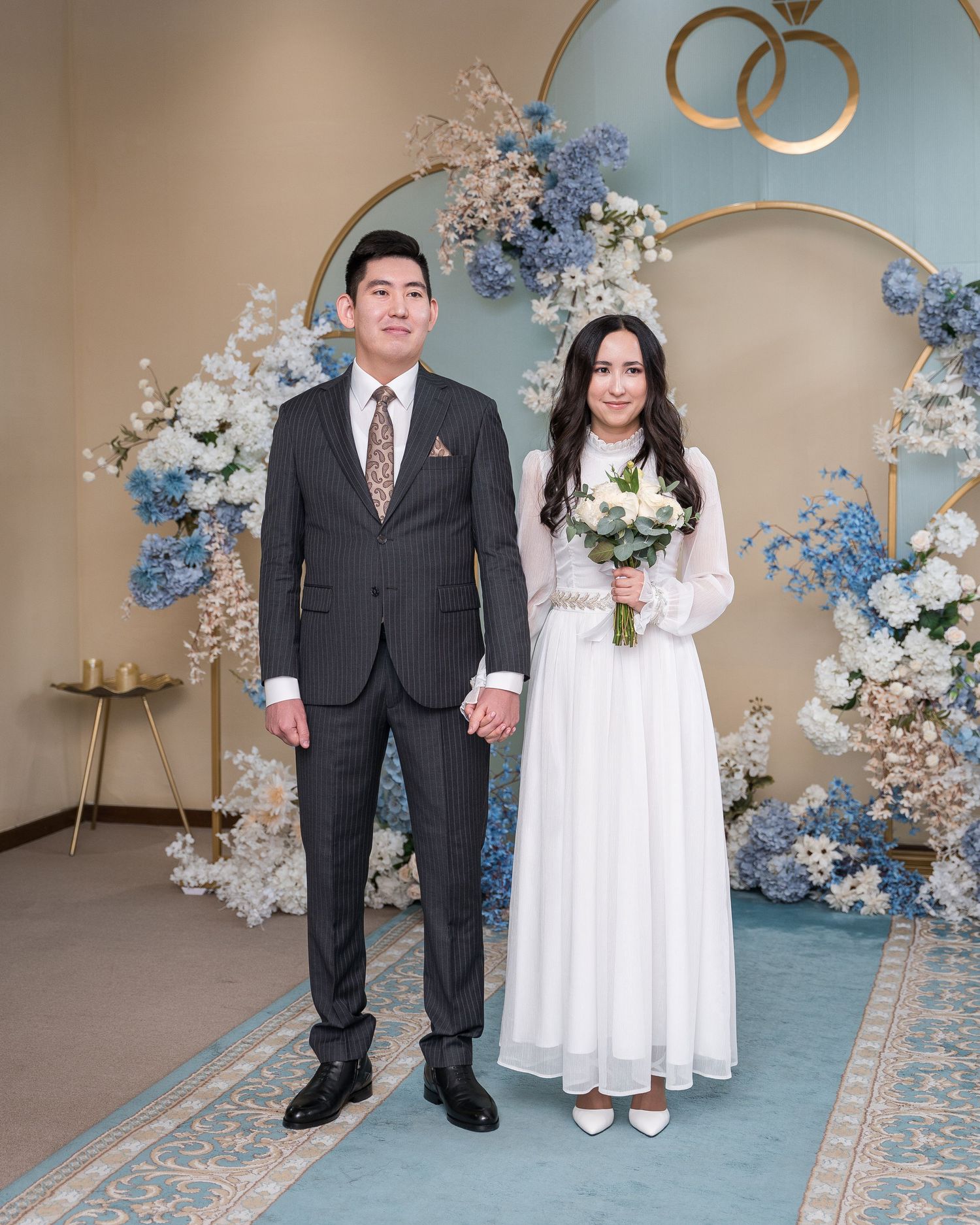 Couple stands hand in hand before blue floral arch with gold rings at elegant wedding ceremony venue.
