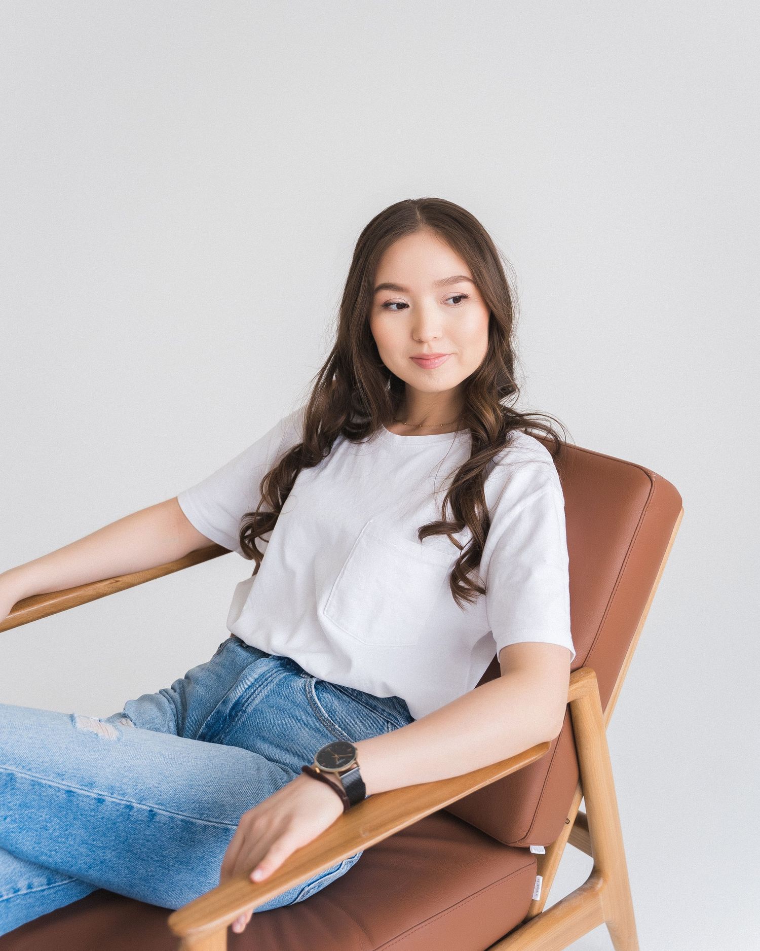 Relaxed poses on brown leather chair in casual white tee and denim.