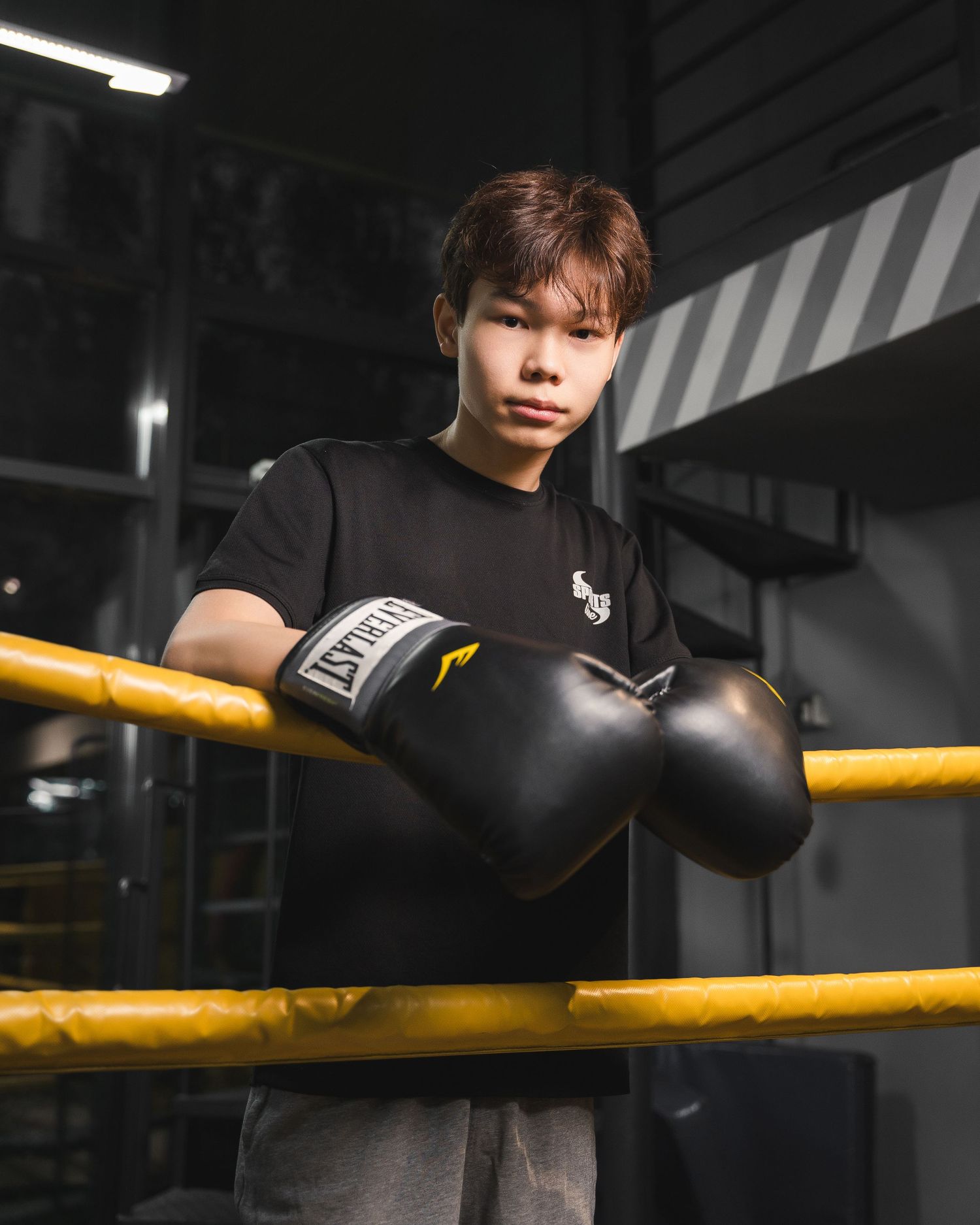 Person wearing boxing gloves leaning against yellow gym railing in dark training space.