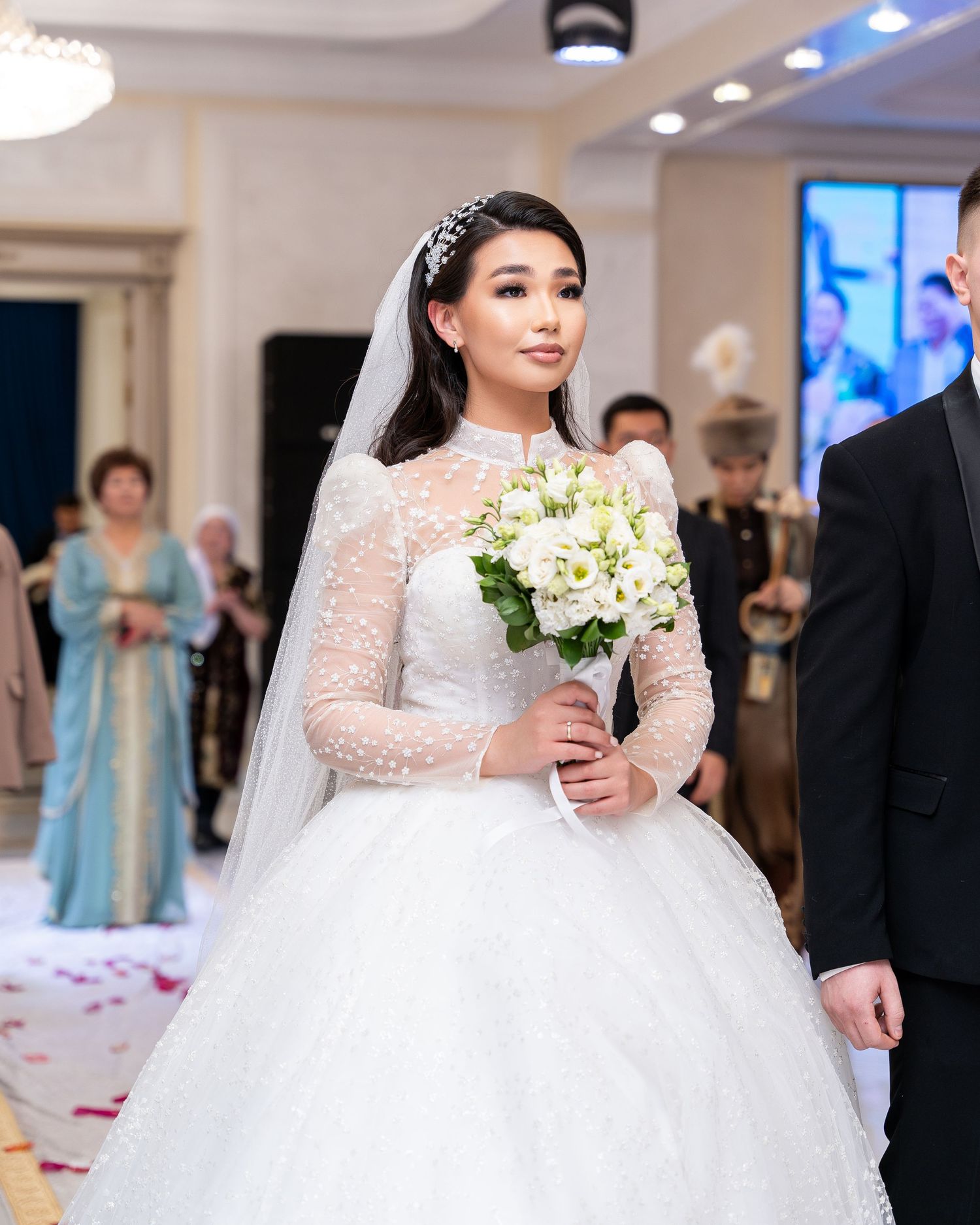 Elegant bride in high-neck lace long-sleeve ballgown carrying white bouquet walking down wedding aisle.