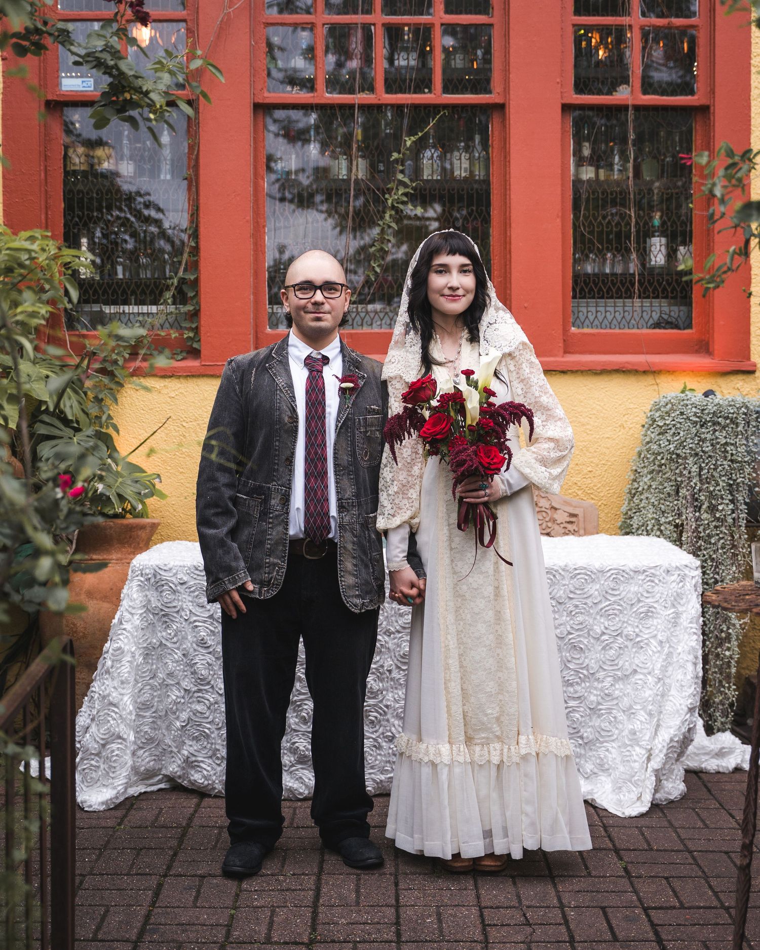 Bride in white lace gown with veil holding red bouquet stands with groom in gray blazer and tie outdoors.