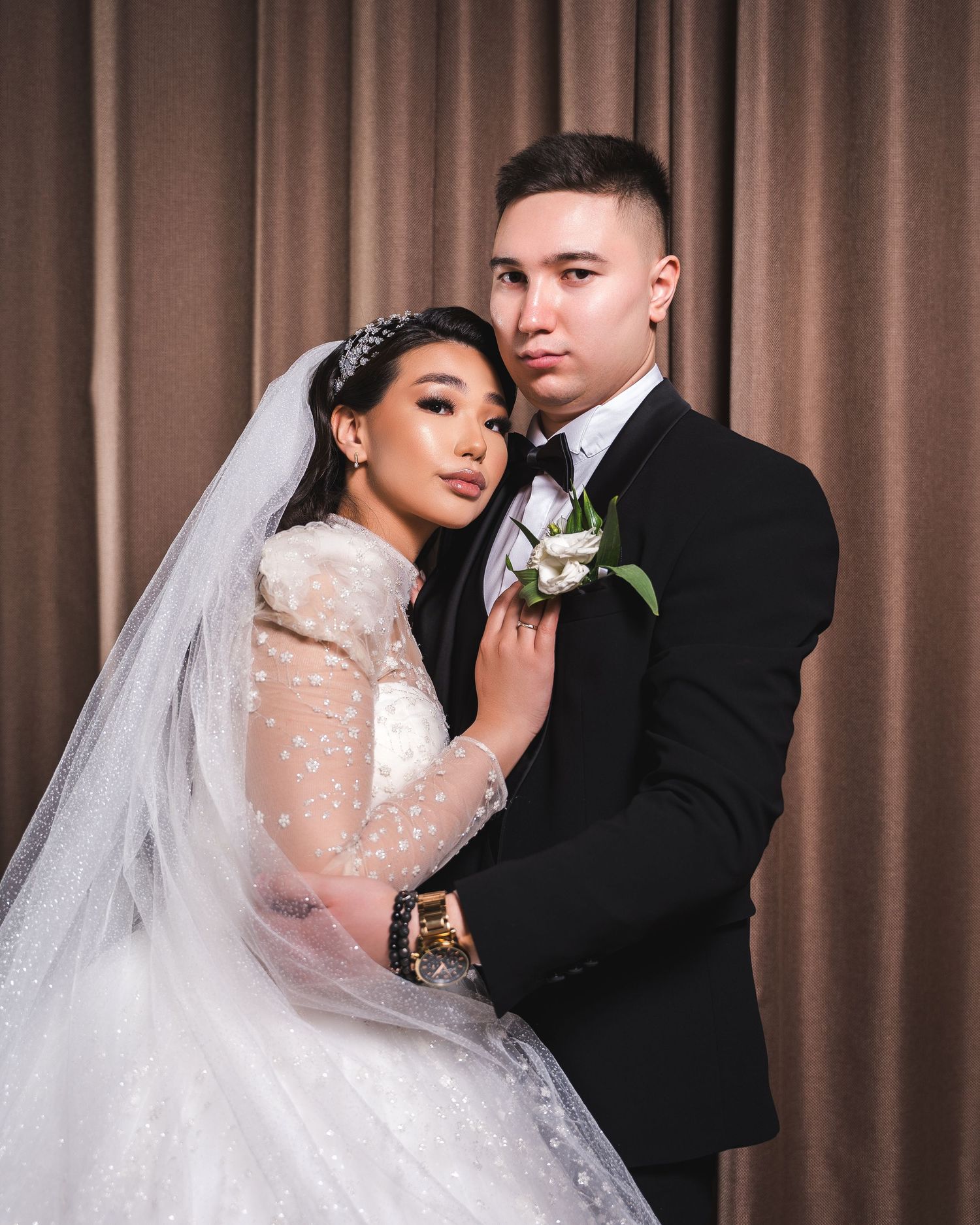 Bride and groom in black tuxedo embracing intimately for formal wedding portrait against beige curtain.