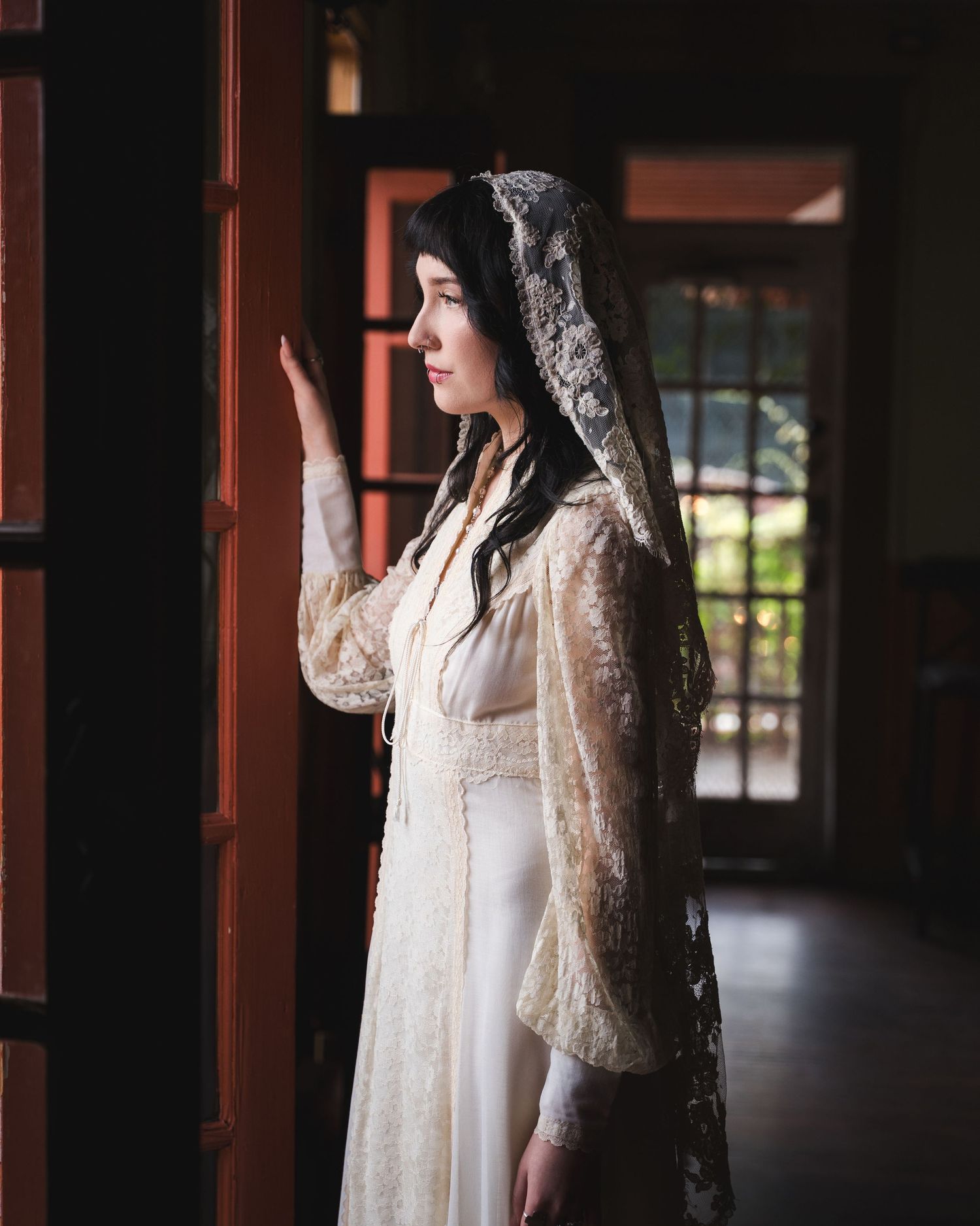 Woman in white lace bridal gown and veil stands by wooden door in soft natural light.