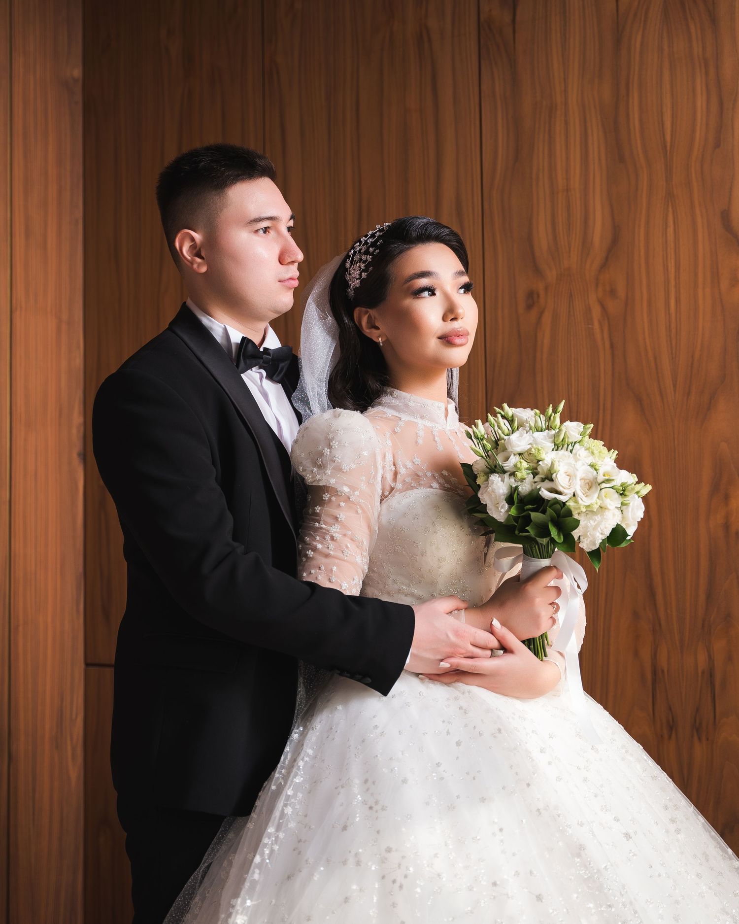 Newlywed couple in tuxedo and lace ballgown holding white bouquet posing against wooden wall backdrop.