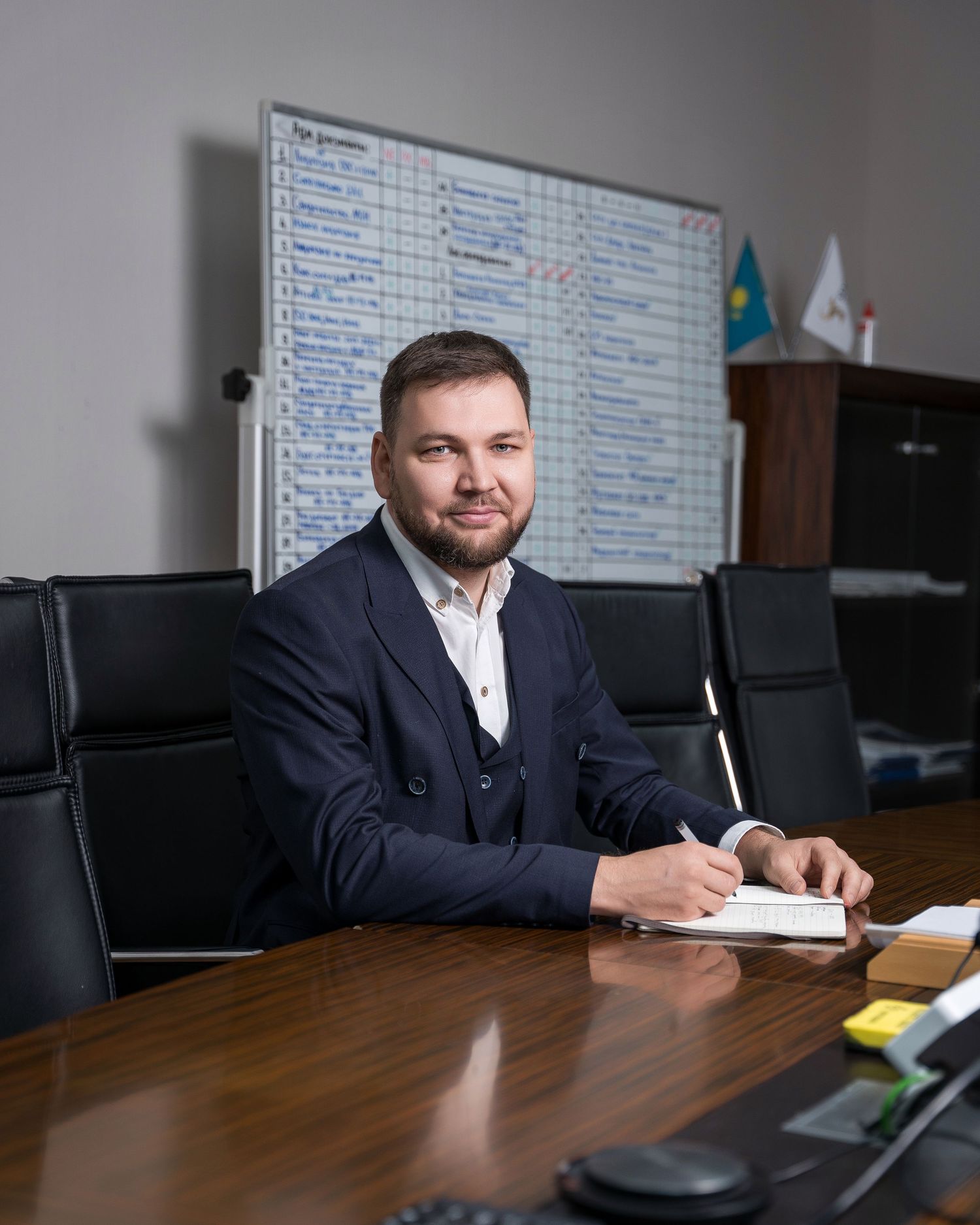Professional in a navy suit seated at a wooden conference table in a modern office setting.