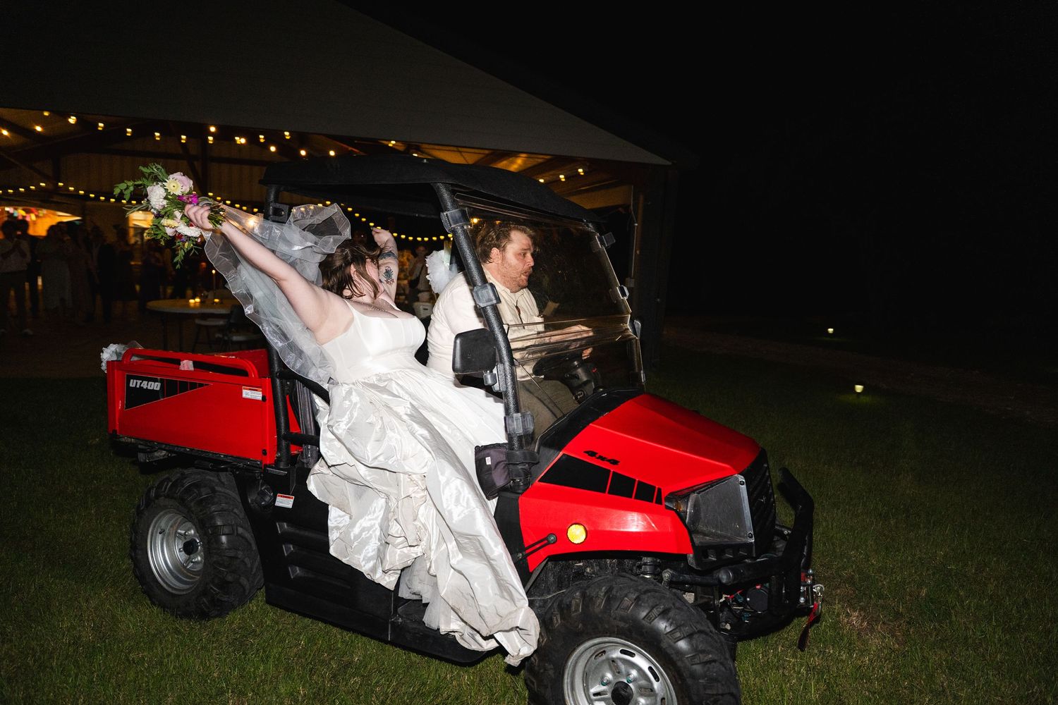 Bride and groom riding a red utility vehicle at night during outdoor wedding reception.