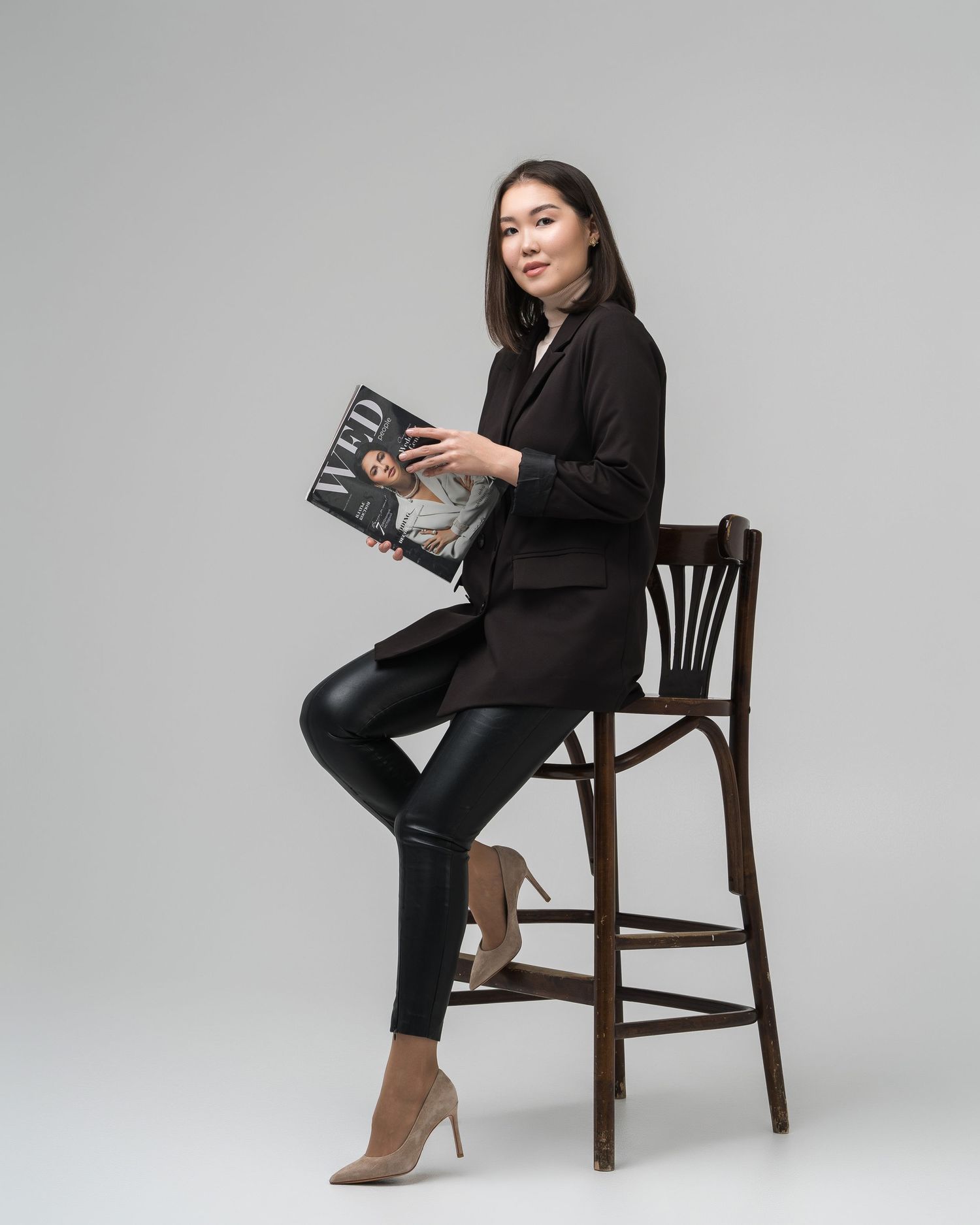 Model in all-black outfit poses elegantly on wooden chair against grey backdrop.
