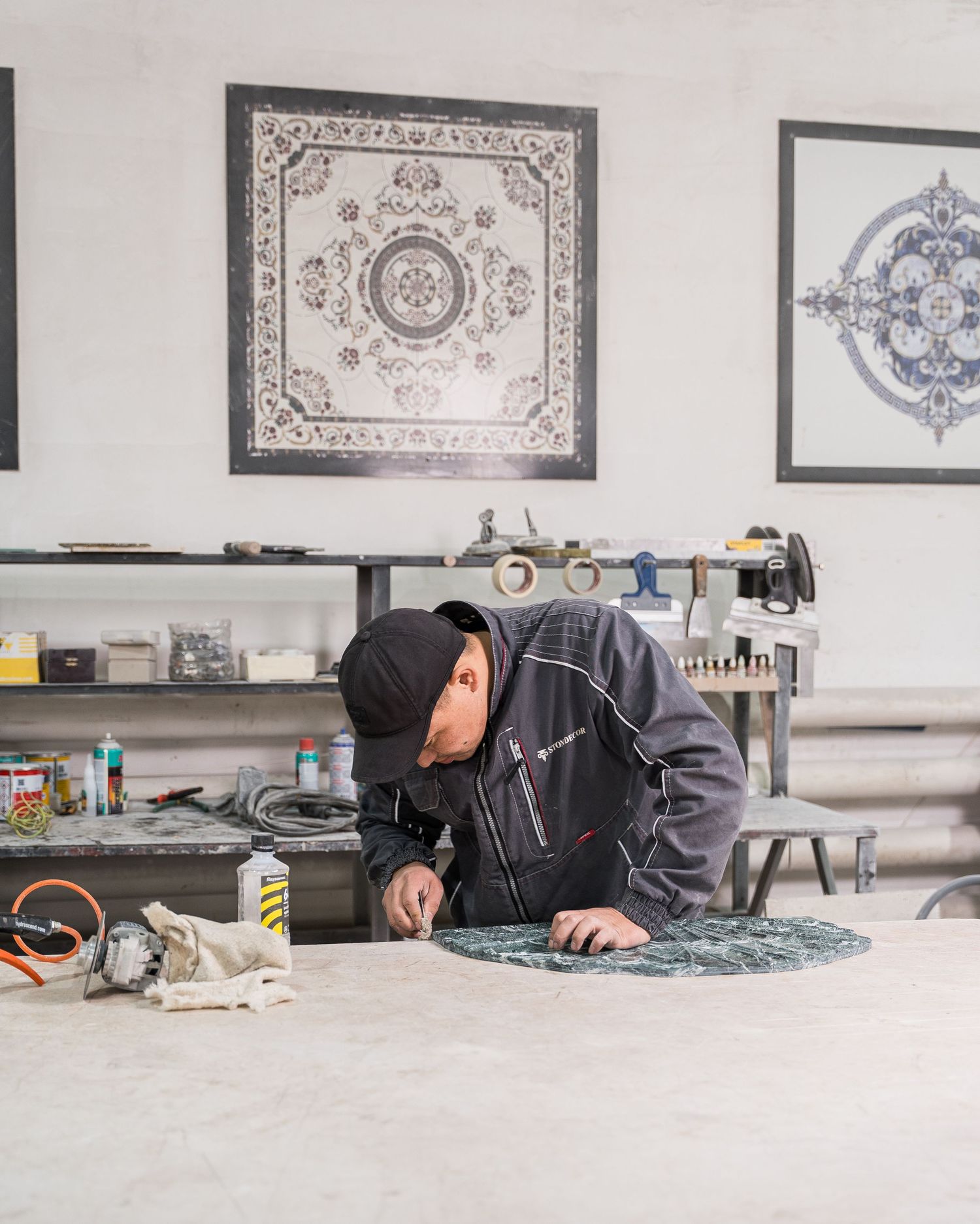 Artisan inspecting intricate stone carving patterns on a workbench with framed designs on wall.