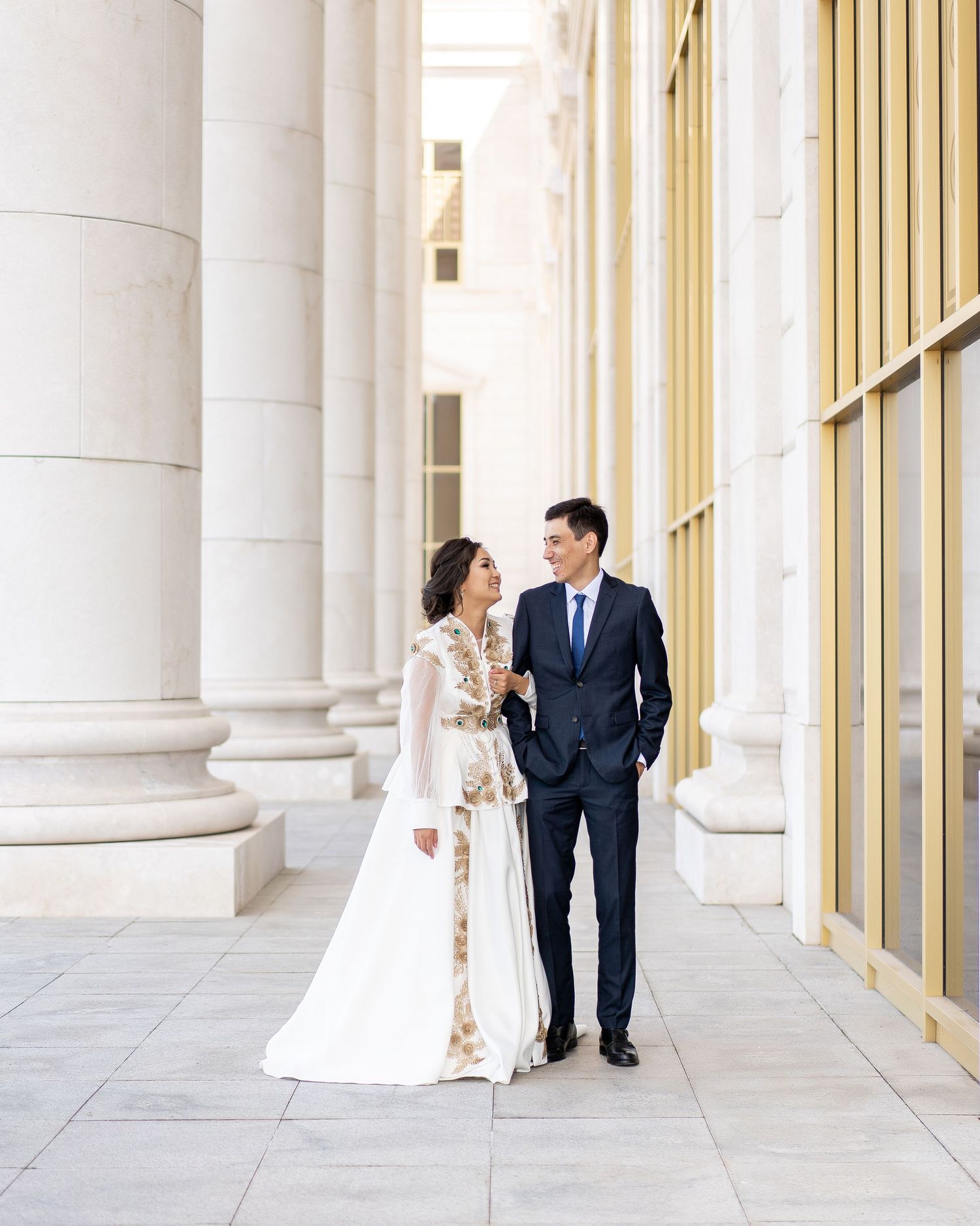 Couple in traditional Kazakh wedding attire walking between grand columns, bride in white and gold dress, groom in navy suit.