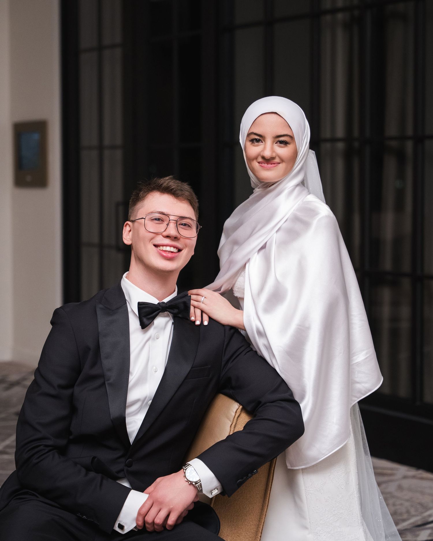 A groom in a black tuxedo and bride in a white hijab and gown pose together for a wedding portrait.
